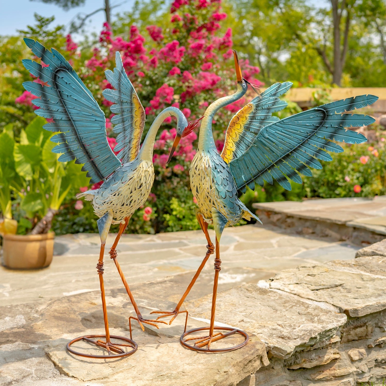 Two decorative metal heron garden statues with spread wings and long orange legs, displayed on a stone patio in a landscaped garden with pink flowers and green foliage in the background.