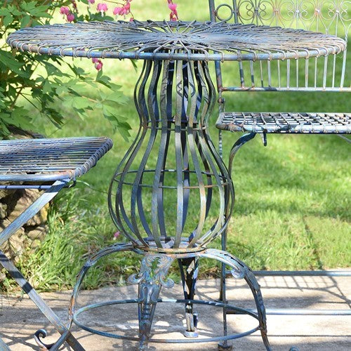 A round metal garden table with a decorative slatted pedestal base, shown outdoors on a patio with grass and plants in the background.