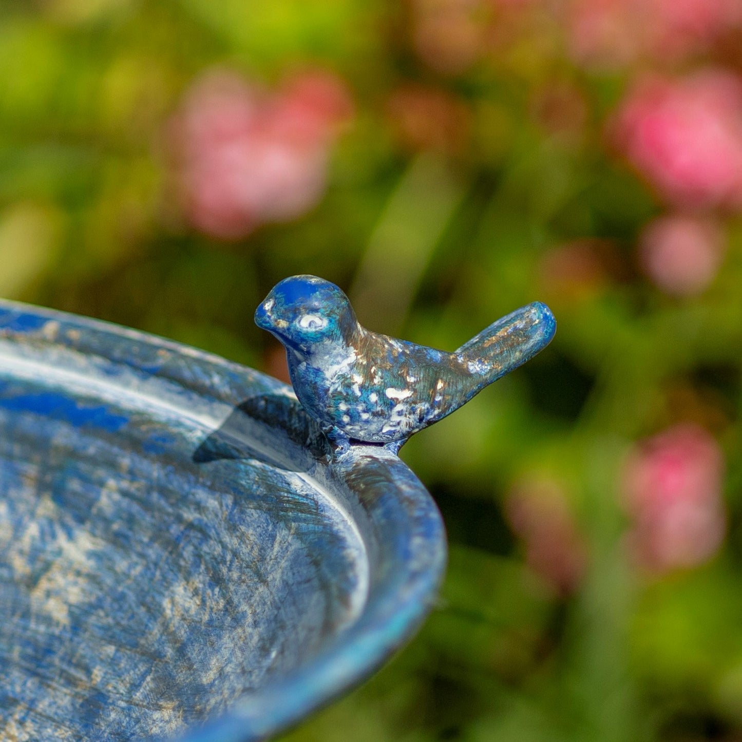 A small blue bird figurine perched on the rim of a blue metal birdbath, photographed outdoors with a blurred green and pink garden background.