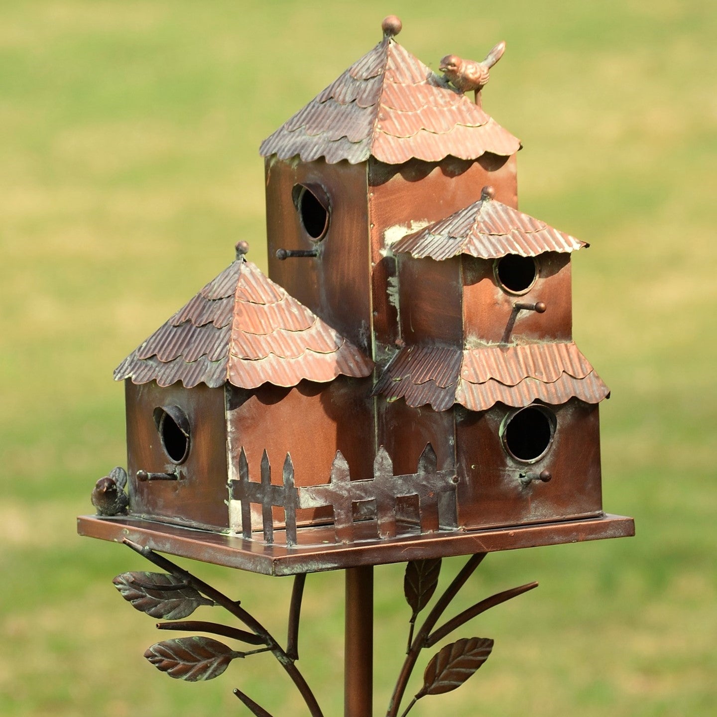 A decorative metal birdhouse set with multiple tiered roofs and small bird perches, mounted on a leaf-shaped metal stand against a blurred green grassy background.