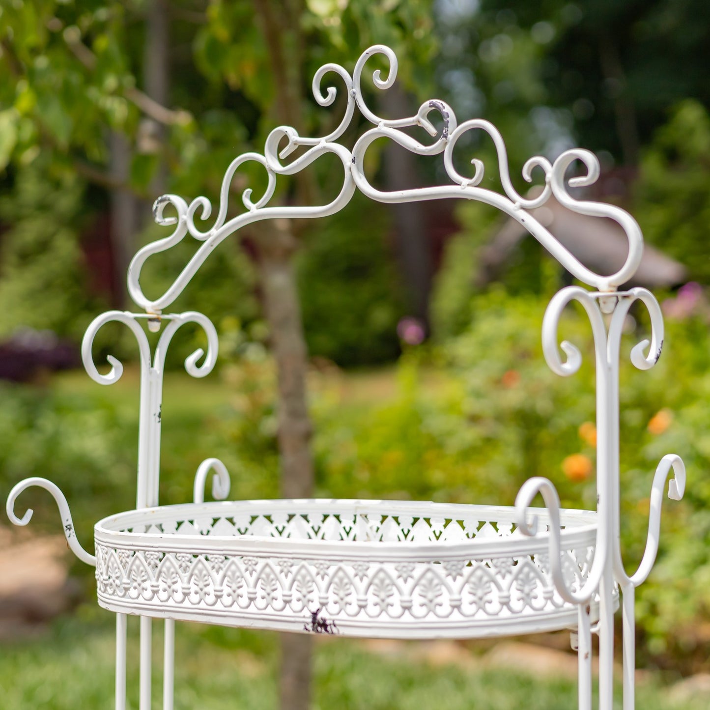 A white ornate metal garden planter stand with a decorative arched top and scalloped front trim, photographed outdoors against a blurred green garden background.