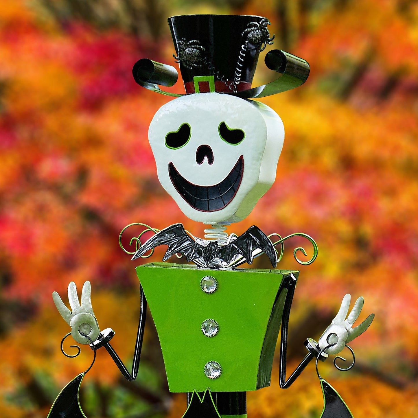 A Halloween metal garden statue styled like a skeleton in a black top hat and green suit, with white gloves and a black bat-wing collar, set against a blurred autumn leaf background.