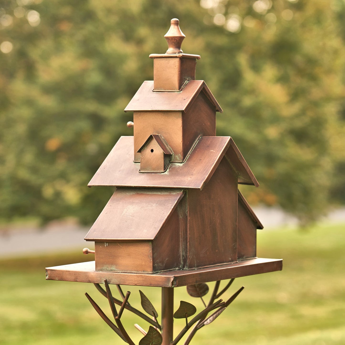 A rustic copper-toned metal birdhouse mounted on a decorative metal tree stake, photographed outdoors against a blurred green garden background.