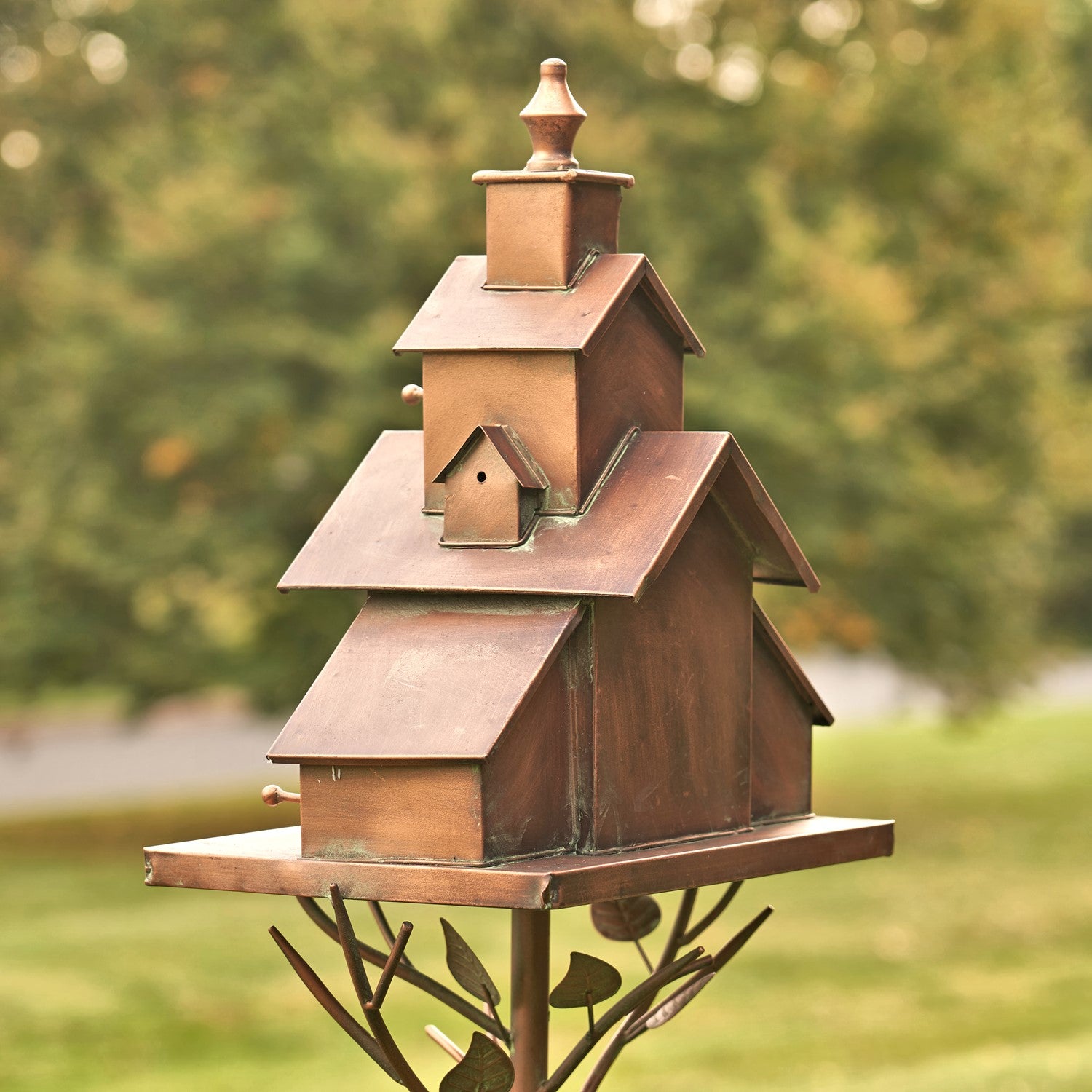 A rustic copper-toned metal birdhouse mounted on a decorative metal tree stake, photographed outdoors against a blurred green garden background.