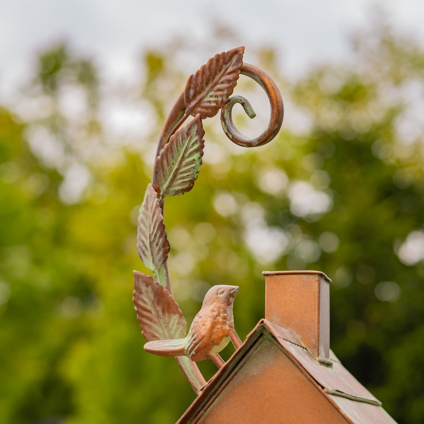 Decorative copper birdhouse with a small bird on top, set against a garden background with flowers and trees