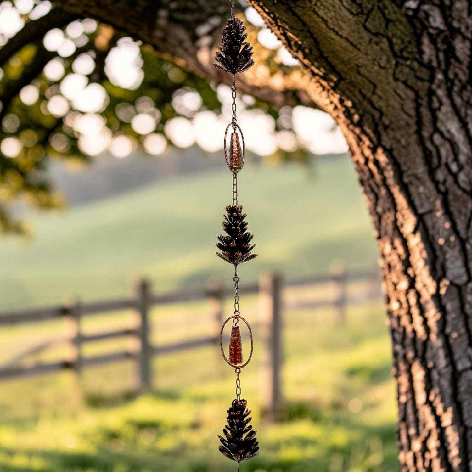 A hanging wind spinner ornament with three pinecones and copper-colored metal accents, suspended from a tree branch with a blurred outdoor landscape in the background.
