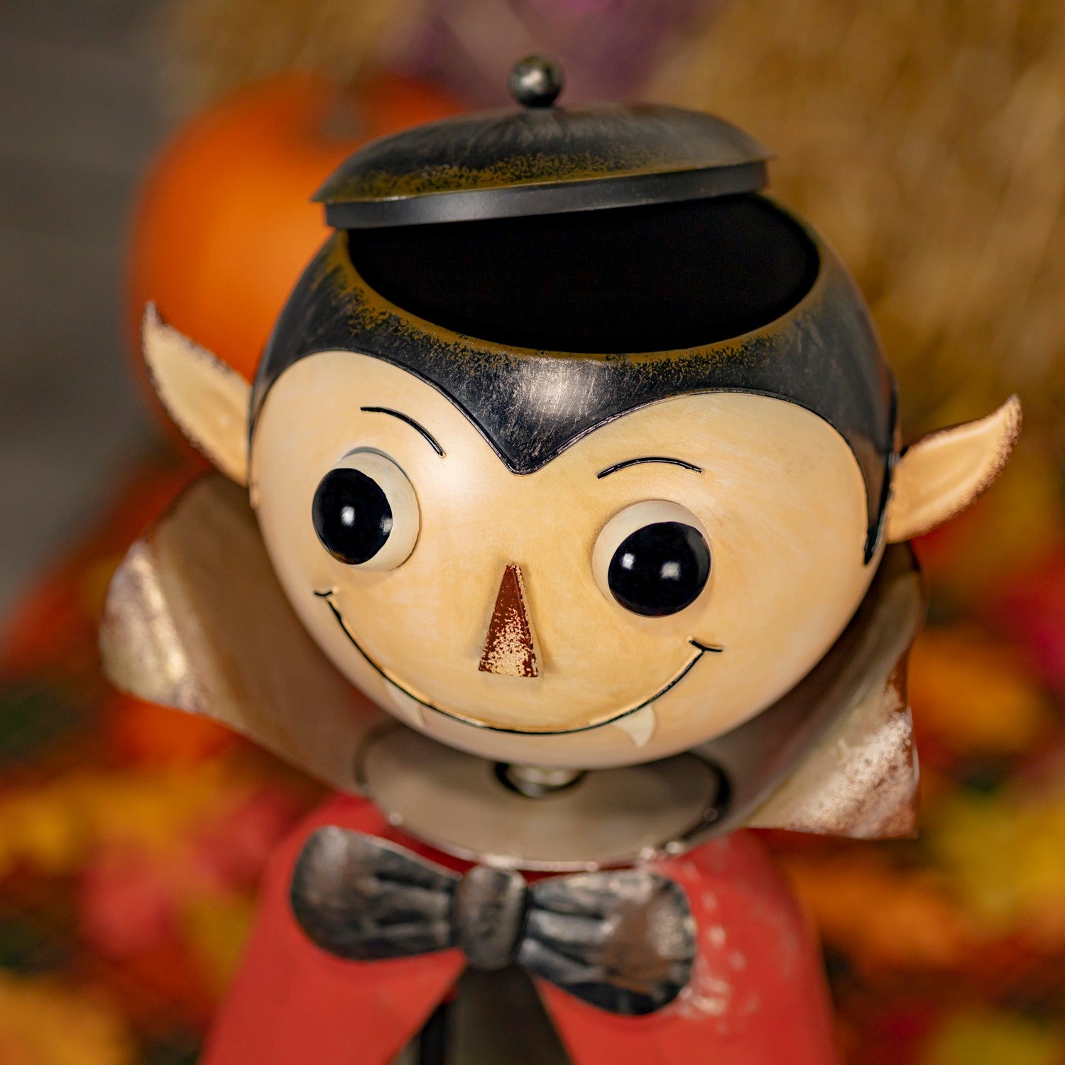 A close-up of a Halloween vampire figurine with a beige face, black hair, pointed ears, a red cape, and a black bow tie, set against a blurred autumn background.