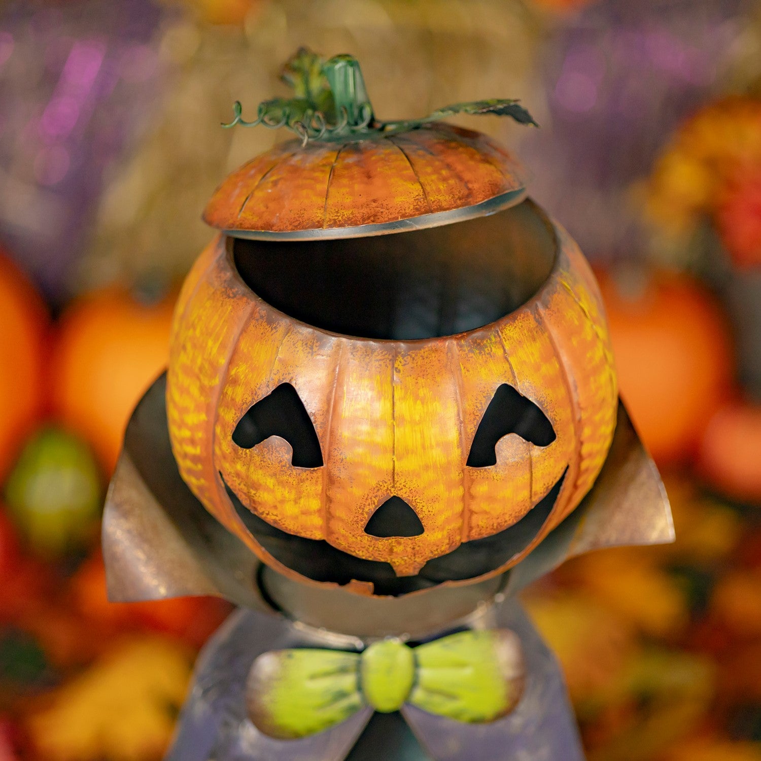 A carved jack-o'-lantern pumpkin container with a lid, styled as a vampire with pointed ears and a green bow tie, set against a blurred autumnal background.