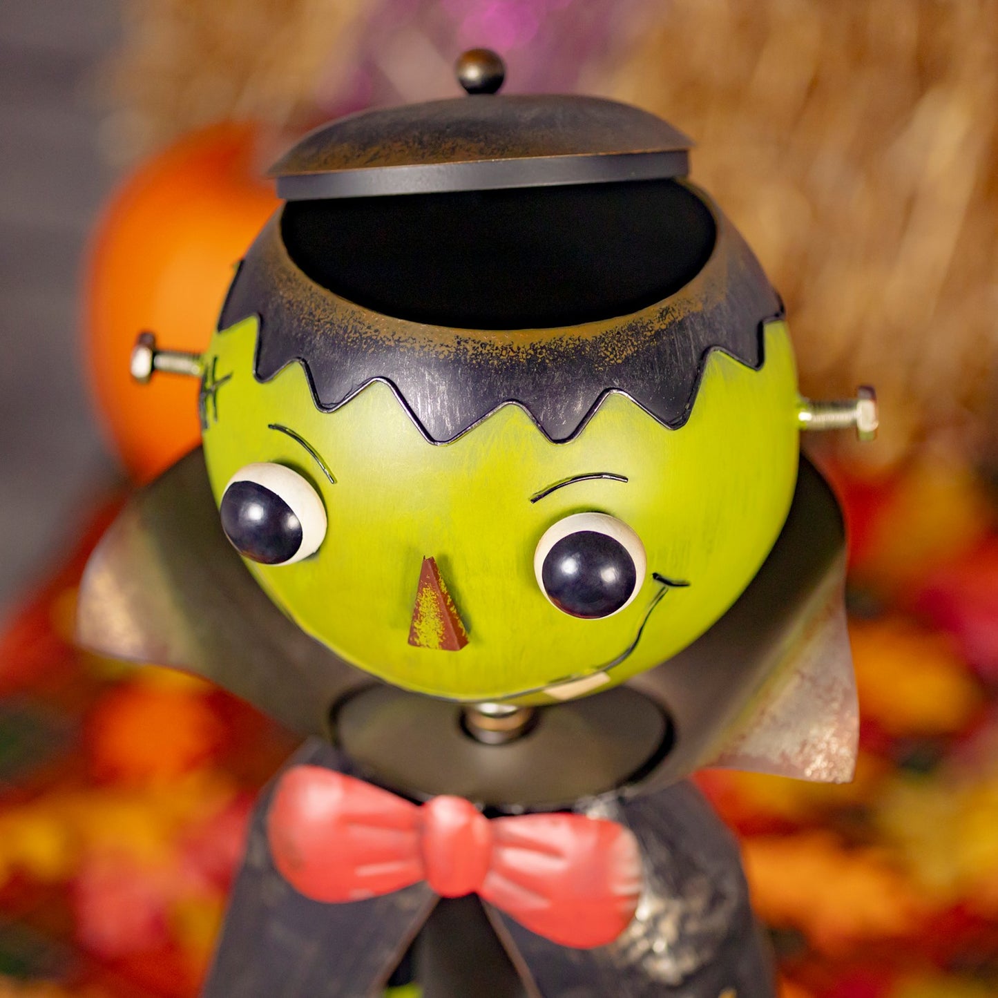 A Frankenstein-themed collectible toy or figurine with a lime green head, black hair, large eyes, a red bow tie, and a dark cape, shown against a blurred autumn background with pumpkins and leaves.