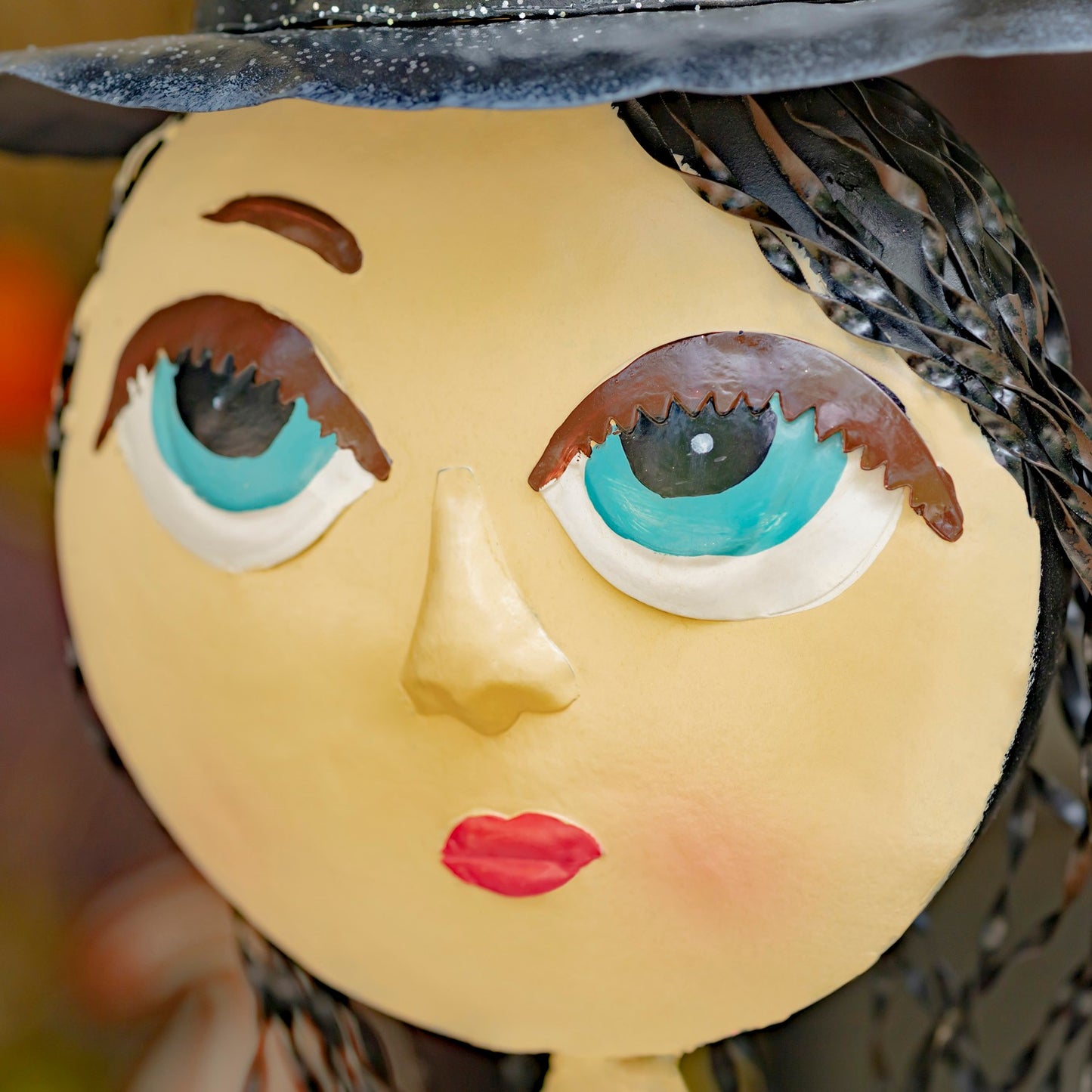 Close-up of a stylized doll or figurine face with a round pale yellow head, large turquoise and white eyes, brown eyebrows and eyelashes, red lips, a black hat, and dark braided hair against a softly blurred background.