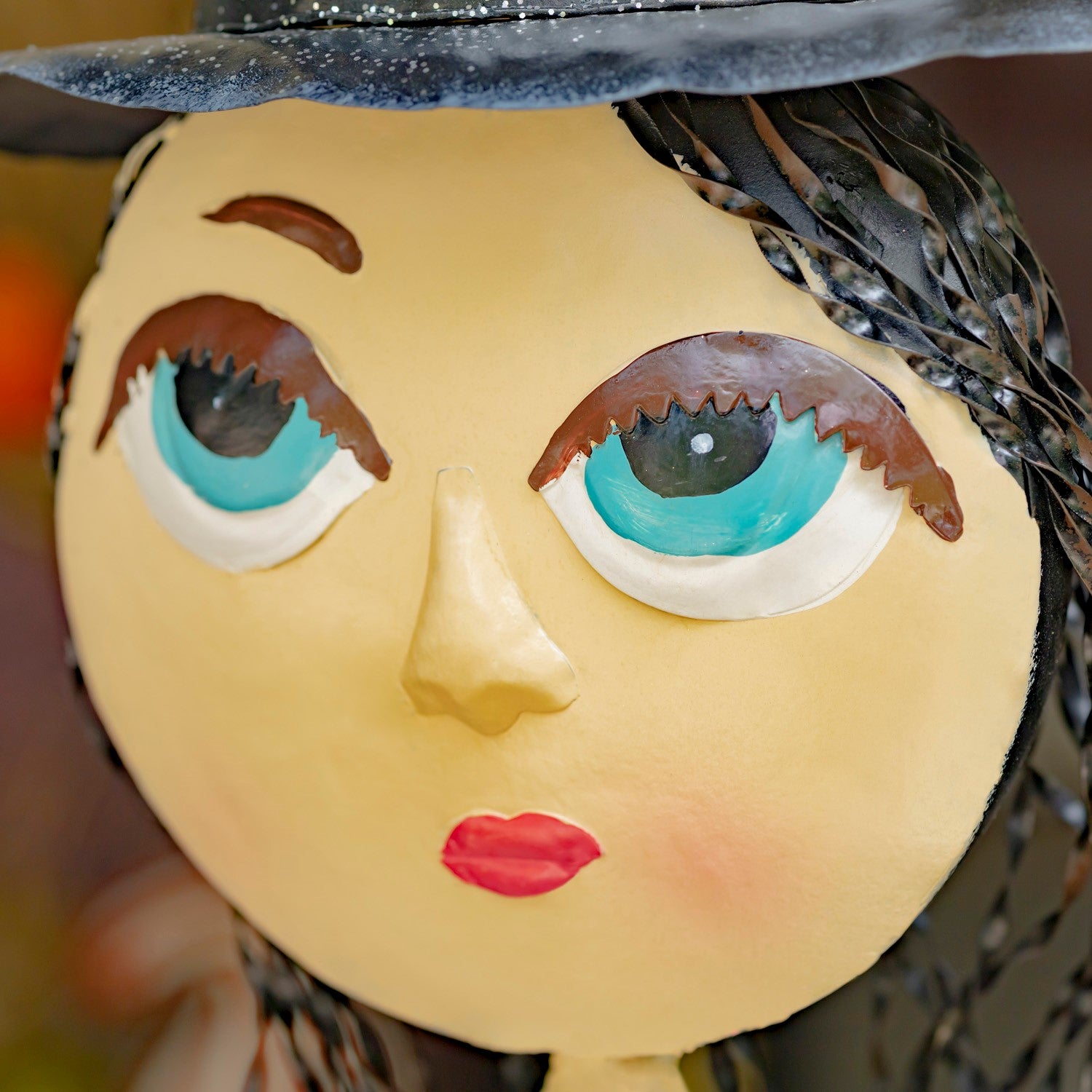 Close-up of a stylized doll or figurine face with a round pale yellow head, large turquoise and white eyes, brown eyebrows and eyelashes, red lips, a black hat, and dark braided hair against a softly blurred background.
