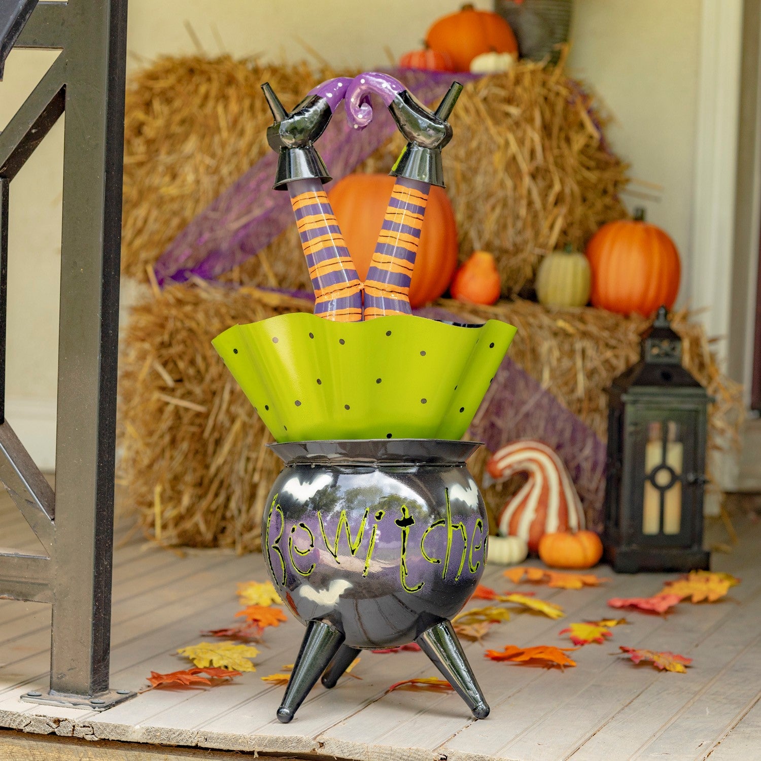 A Halloween decorative witch cauldron set on a porch, featuring a black cauldron with 'Beware the' written on it, a bright green rim, and legs in orange-and-purple striped socks rising out of the cauldron.