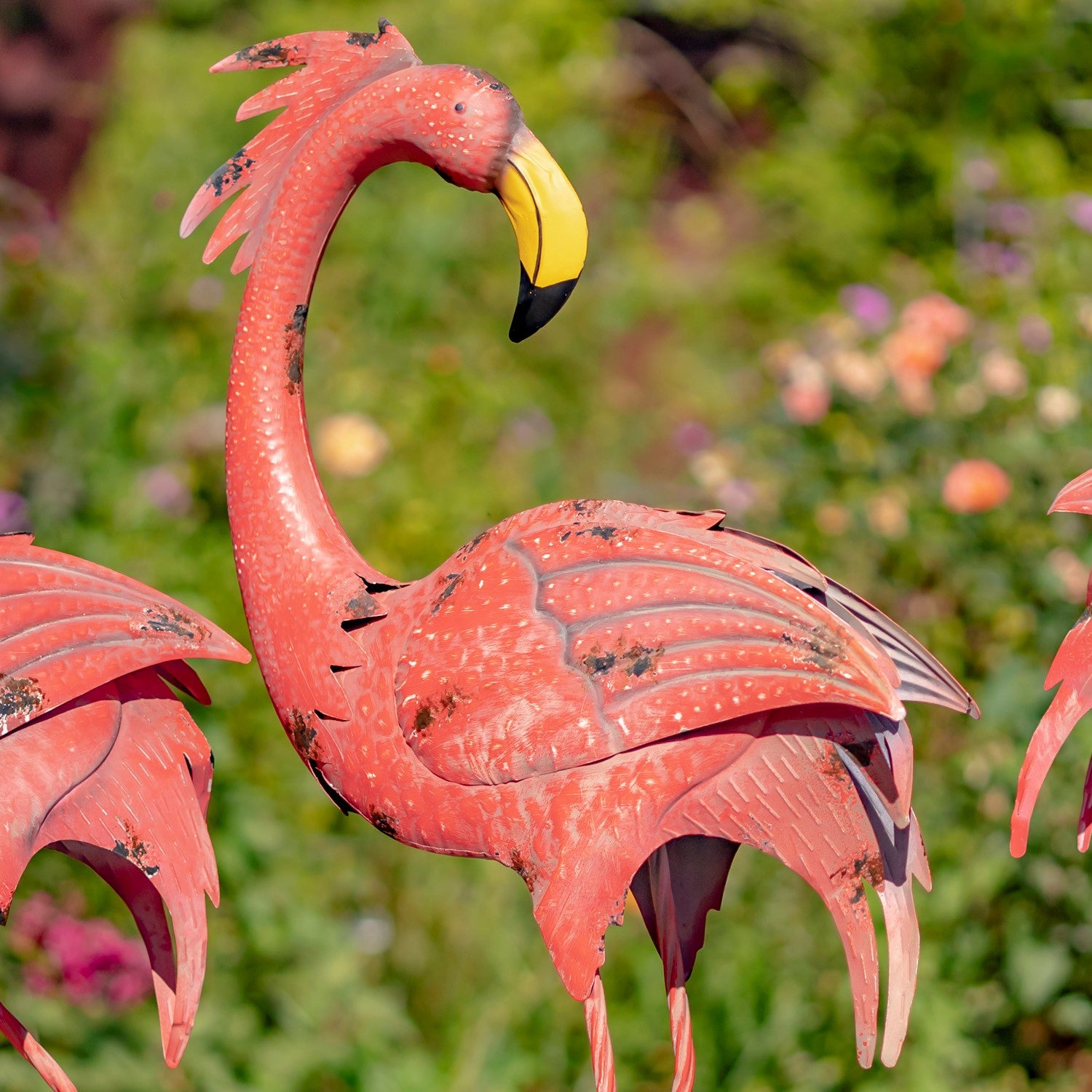 A pink flamingo garden statue with a yellow and black beak, shown in a blurred green outdoor garden setting.