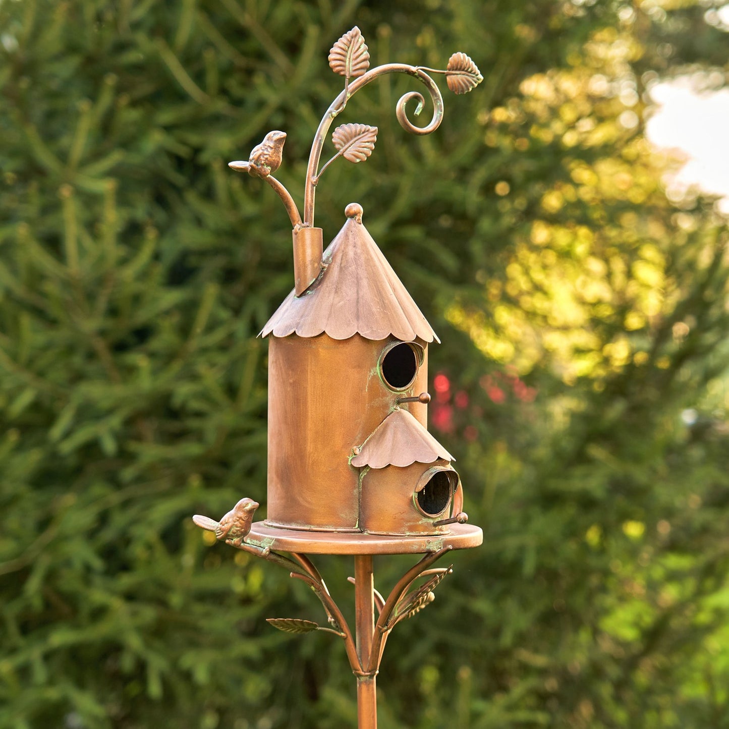 Decorative copper birdhouse with leaf accents on a stand against a blurred green background