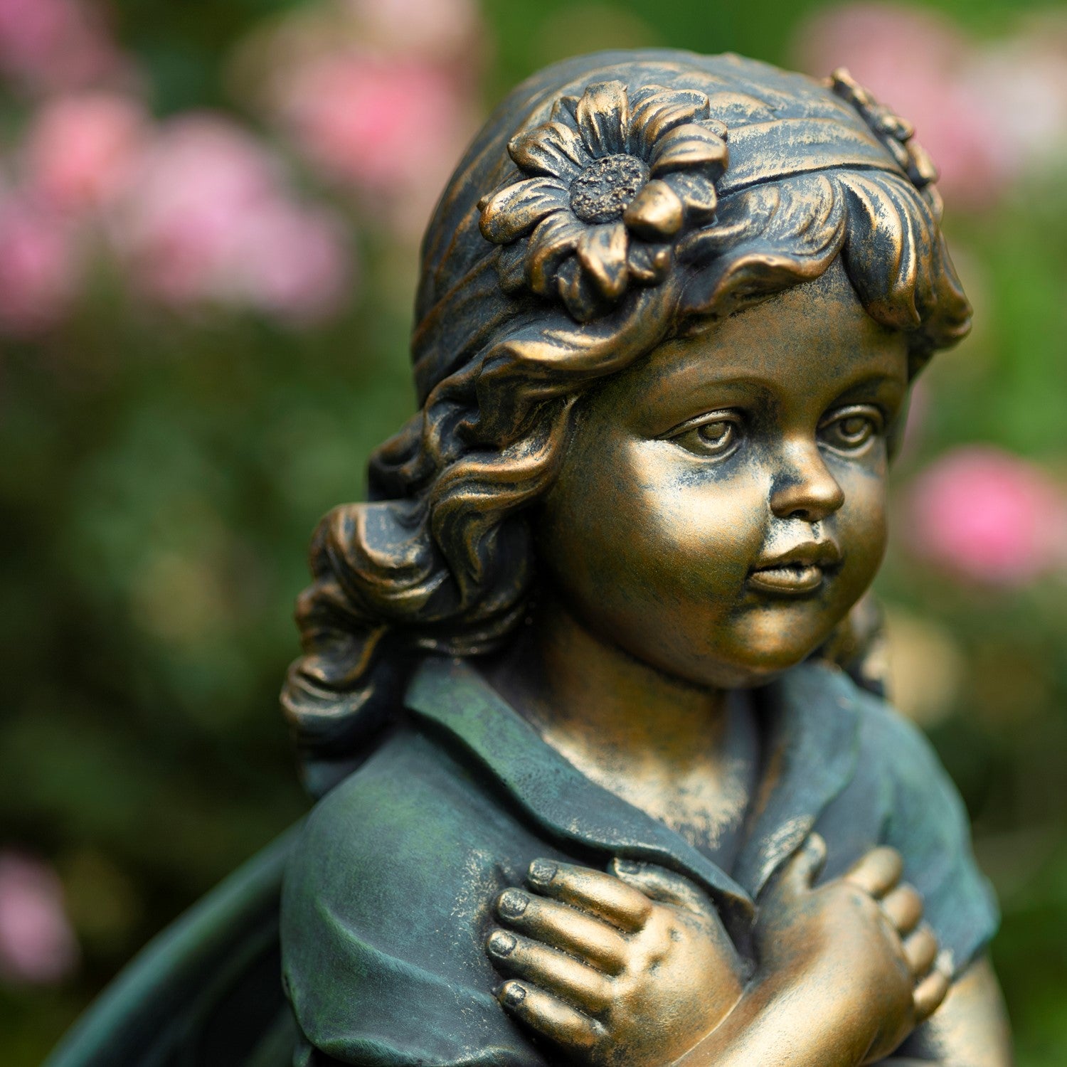 Bronze-toned garden statue of a young girl with a flower in her hair, shown in a close-up outdoor shot with blurred pink flowers and green foliage in the background.