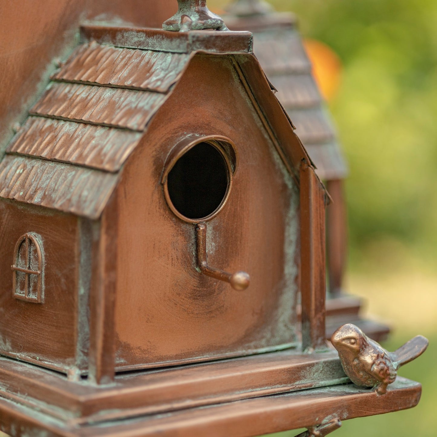 A close-up of a decorative miniature birdhouse in a weathered copper or bronze finish with a small bird figurine on the front perch, set against a blurred green outdoor background.