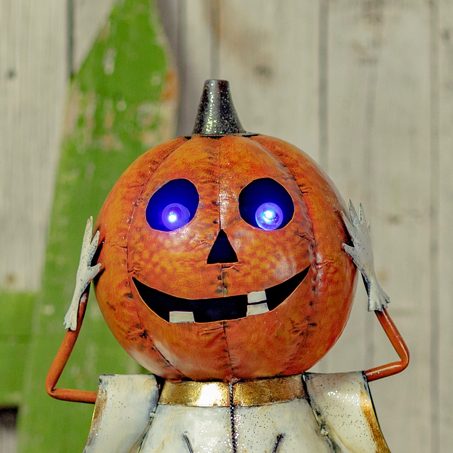 A Halloween metal garden statue of a smiling jack-o'-lantern with glowing blue LED eyes, a silver top hat, and white hands, set against a rustic wooden background.