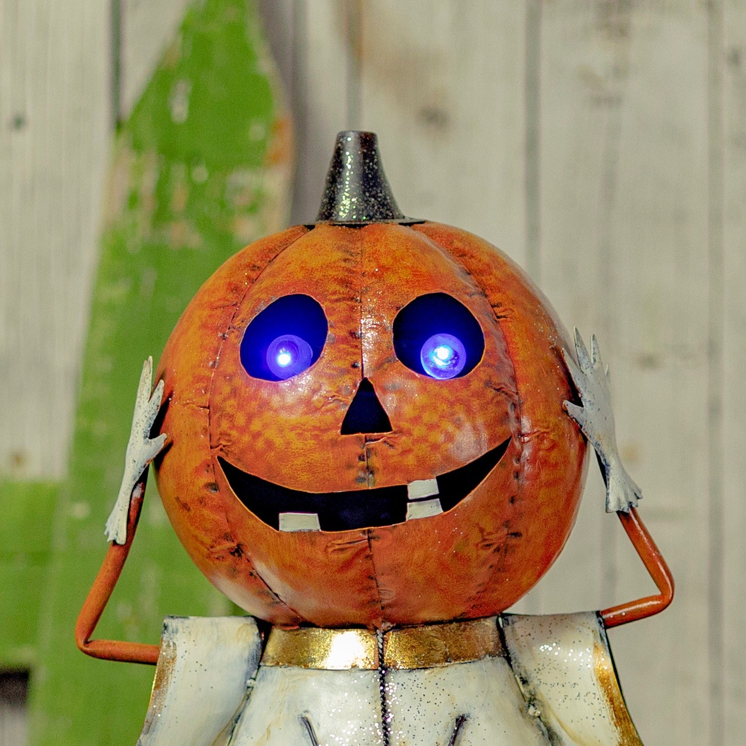 A Halloween metal garden statue of a smiling jack-o'-lantern with glowing blue LED eyes, a silver top hat, and white hands, set against a rustic wooden background.