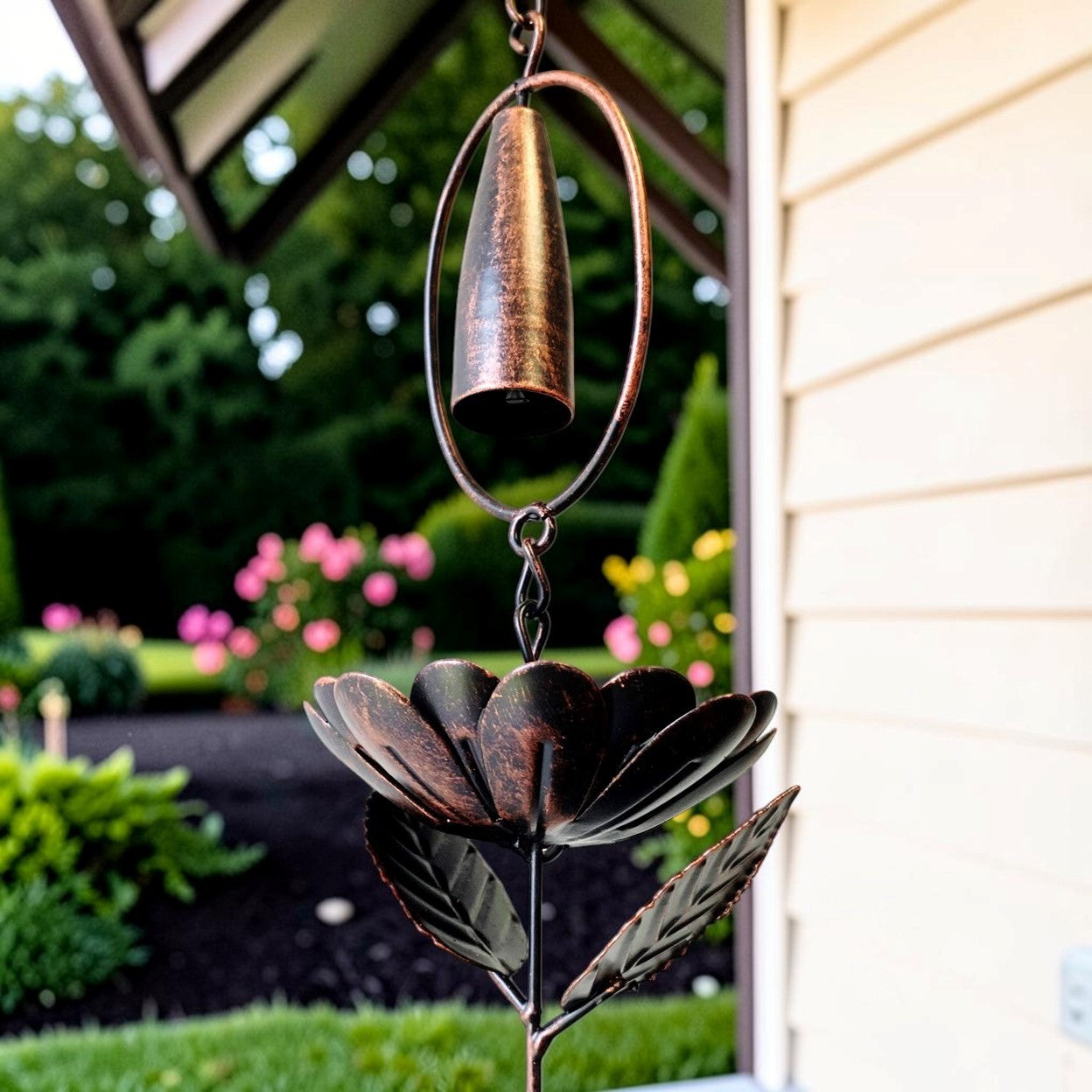 A bronze metal garden wind chime shaped like a flower with a hanging bell and leafy stem, shown outdoors in front of a house with a garden background.