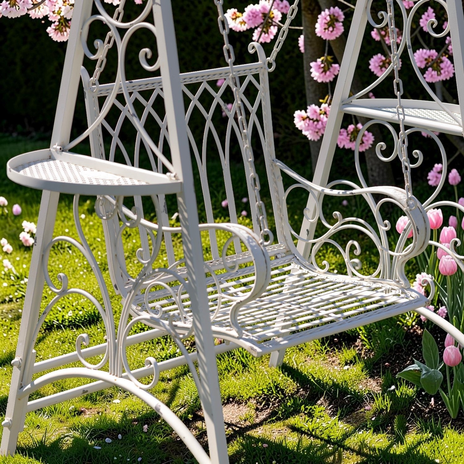 A white wrought iron garden swing bench with decorative scrollwork, set in a grassy garden with pink flowers hanging nearby.