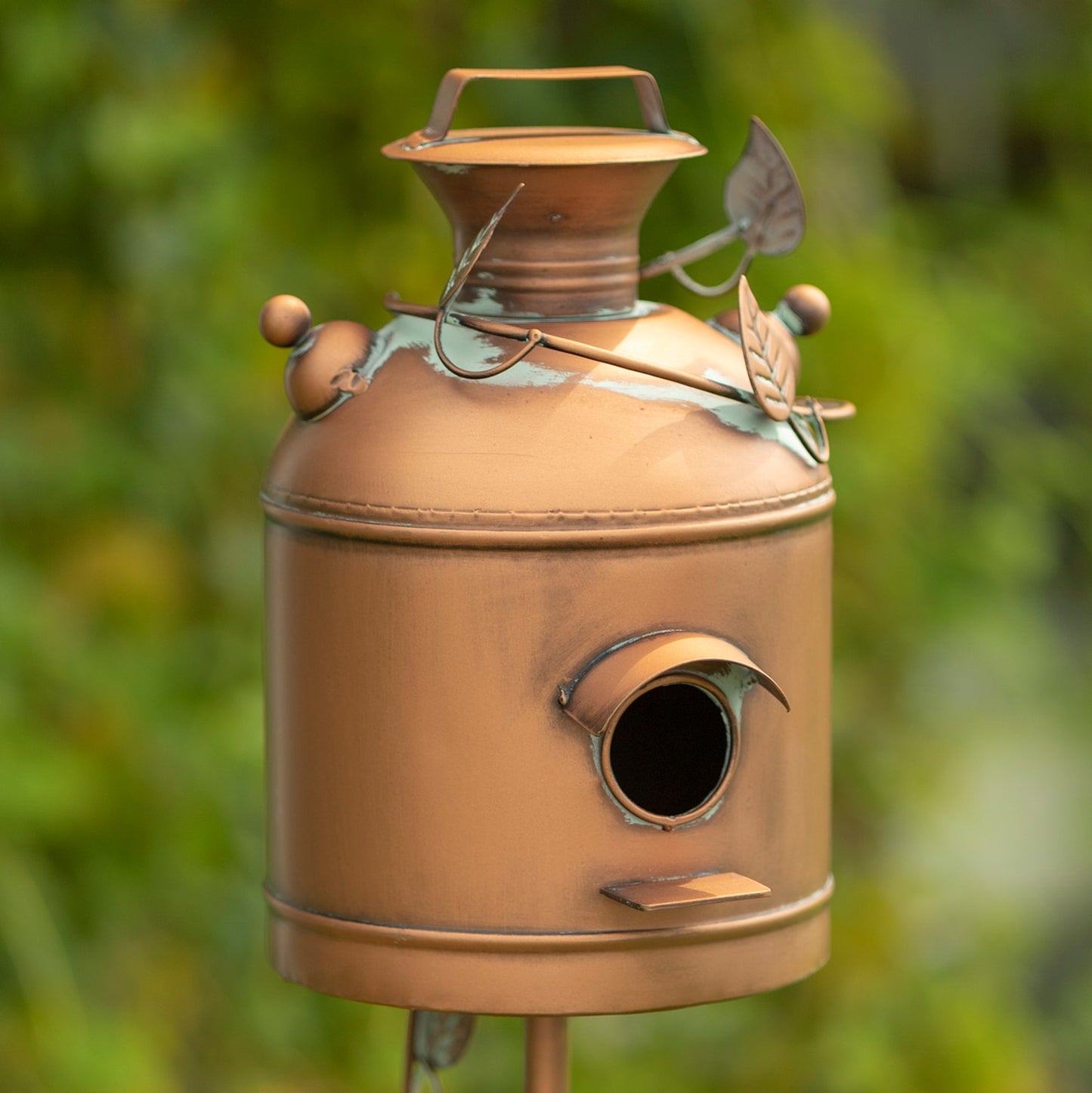 A decorative metal birdhouse shaped like a vintage milk canister, with a copper-toned finish, leaf accents, and a small round entrance hole, photographed outdoors against a blurred green background.
