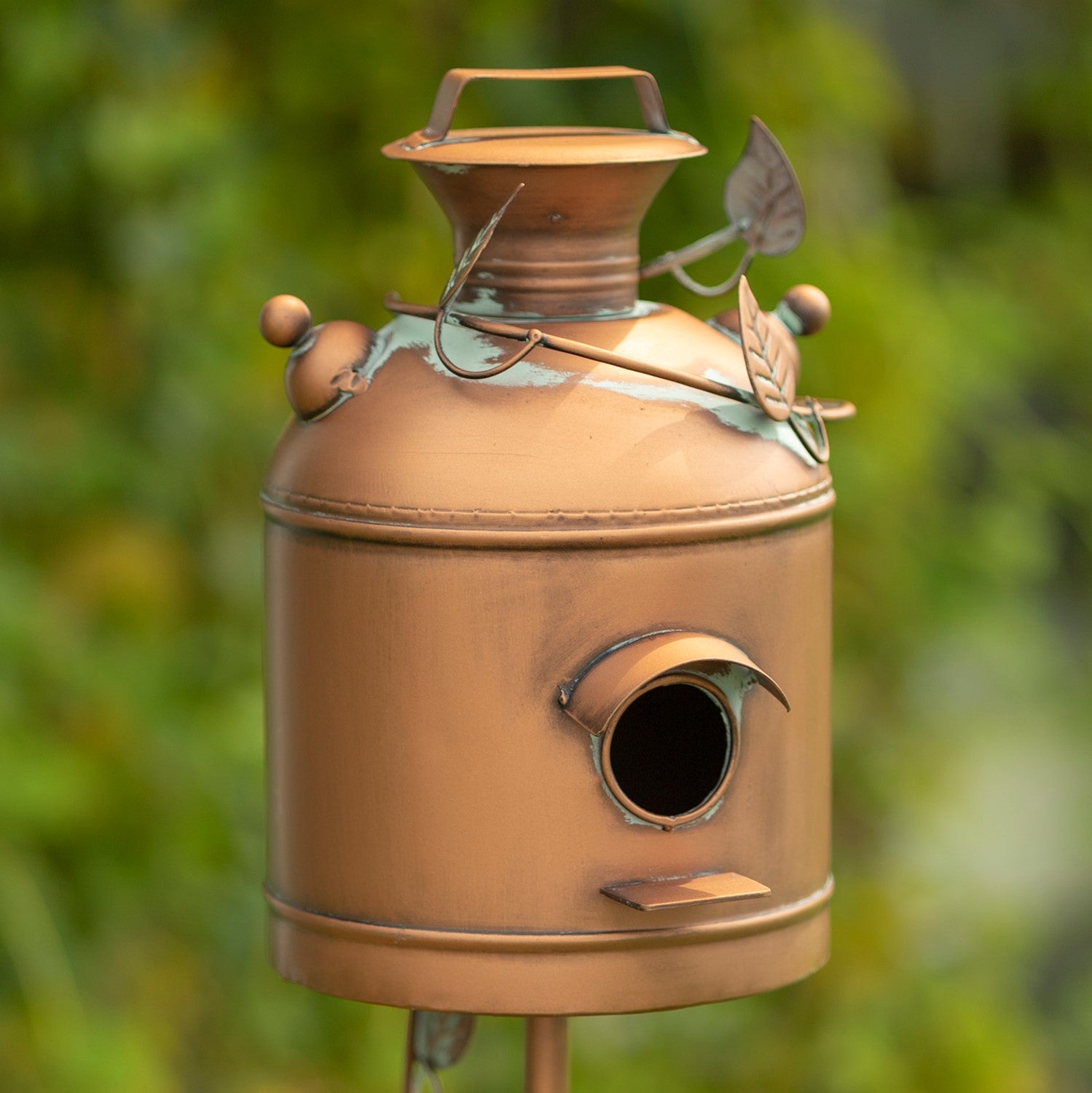 A decorative metal birdhouse shaped like a vintage milk canister, with a copper-toned finish, leaf accents, and a small round entrance hole, photographed outdoors against a blurred green background.