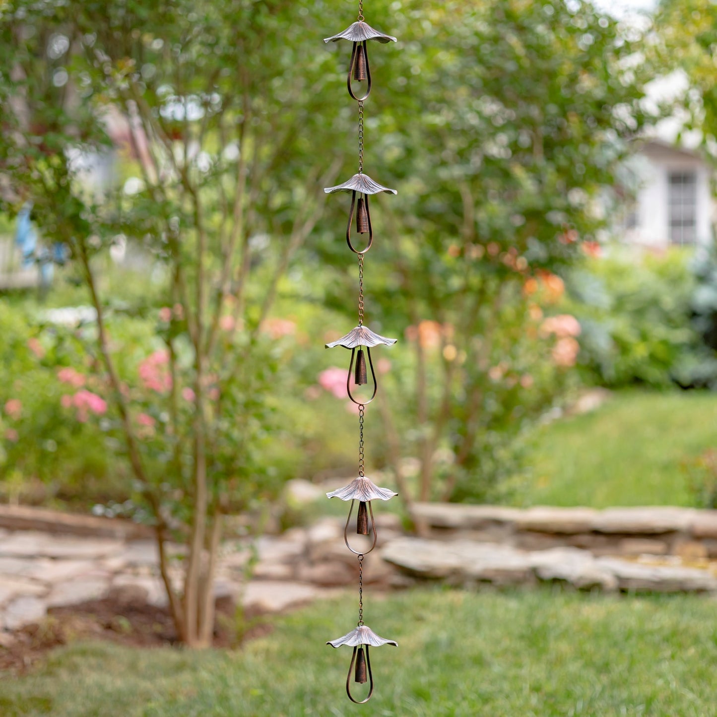 A vertical garden wind spinner with five metallic flower-shaped blades and clear hanging loops, shown outdoors against a blurred green garden background.