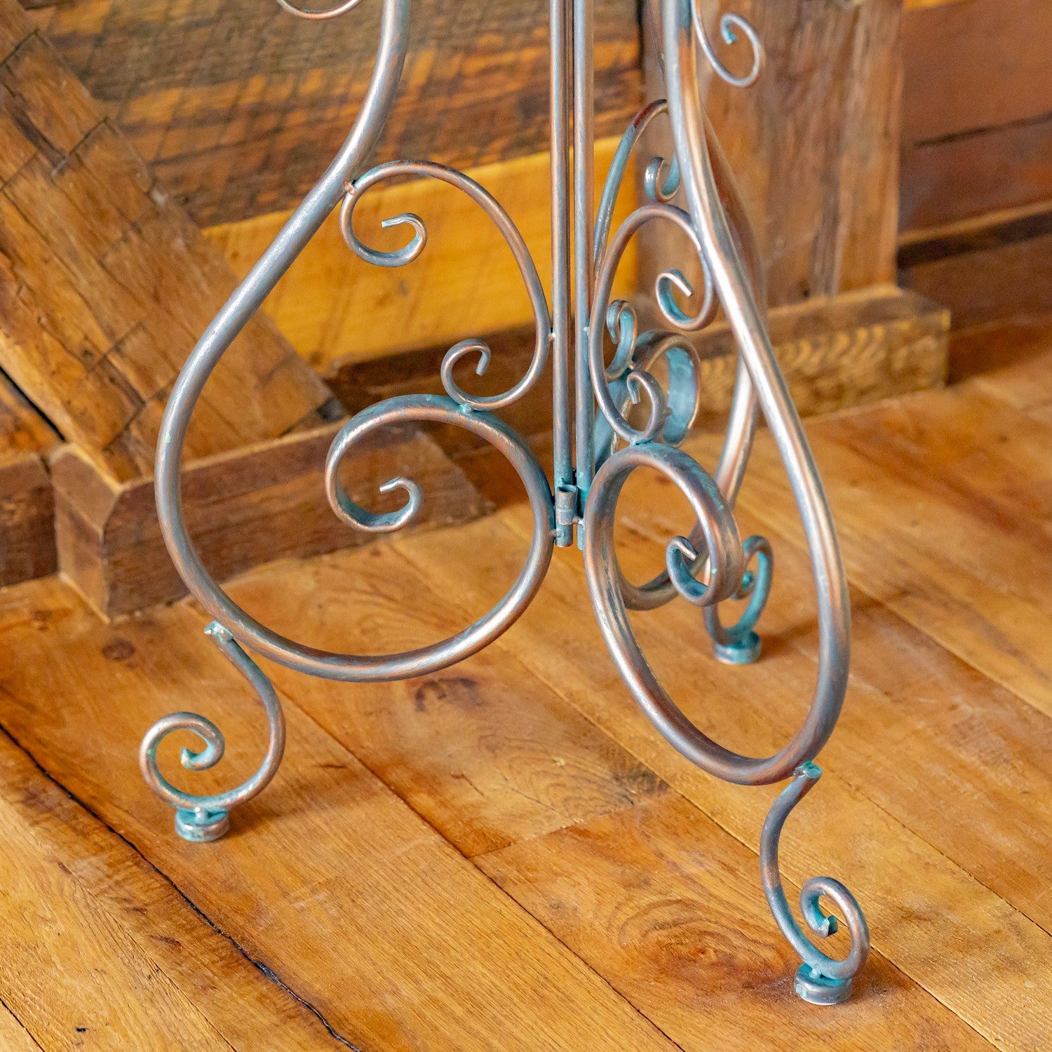 An ornate metal display stand with swirling scrollwork and small feet, finished in a silver-toned metal with a light blue patina, shown on a rustic wooden floor.