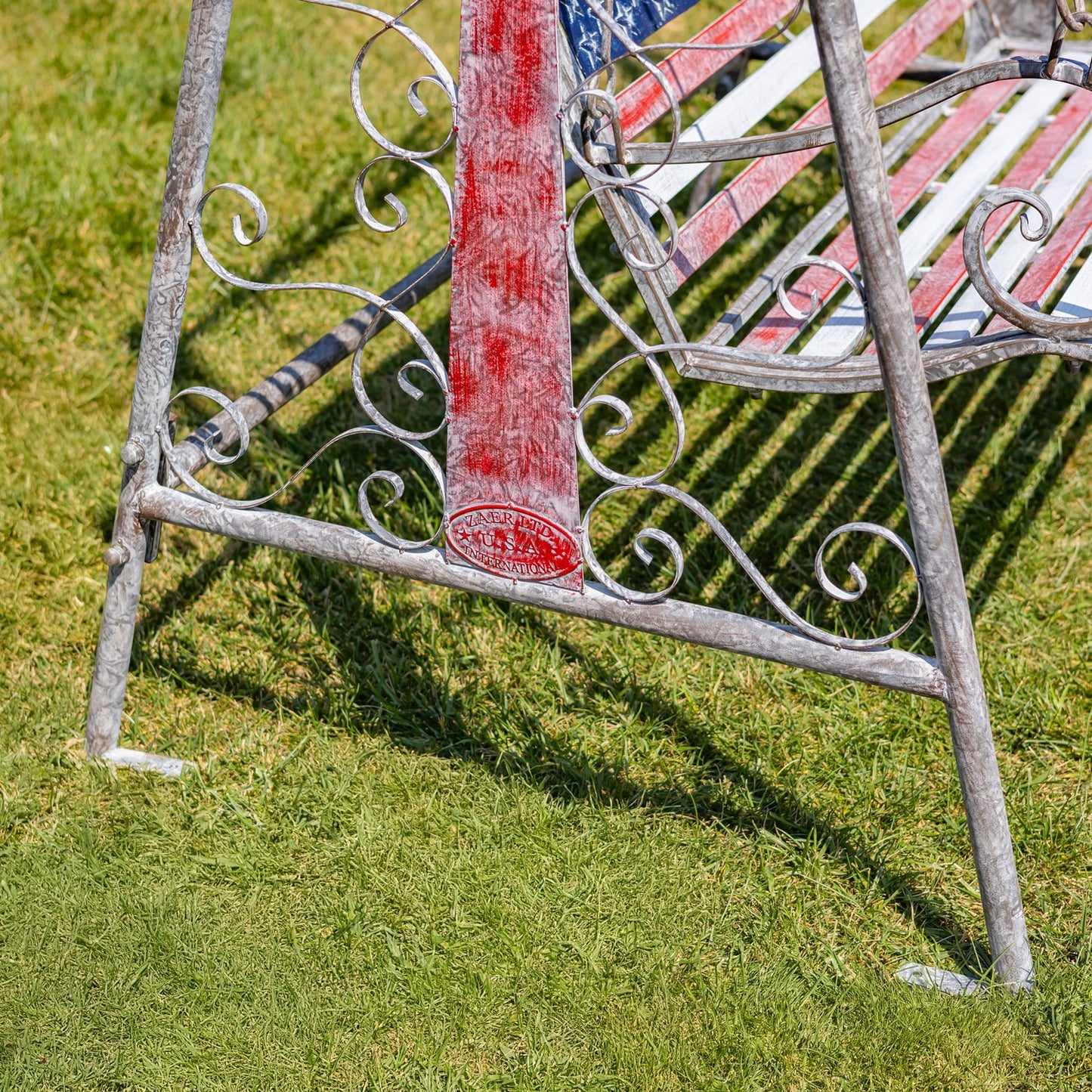 A close-up outdoor shot of a weathered metal garden bench on green grass. The bench has ornate scrollwork on the frame, a distressed red vertical support, and a red, white, and blue striped slatted seat and backrest.
