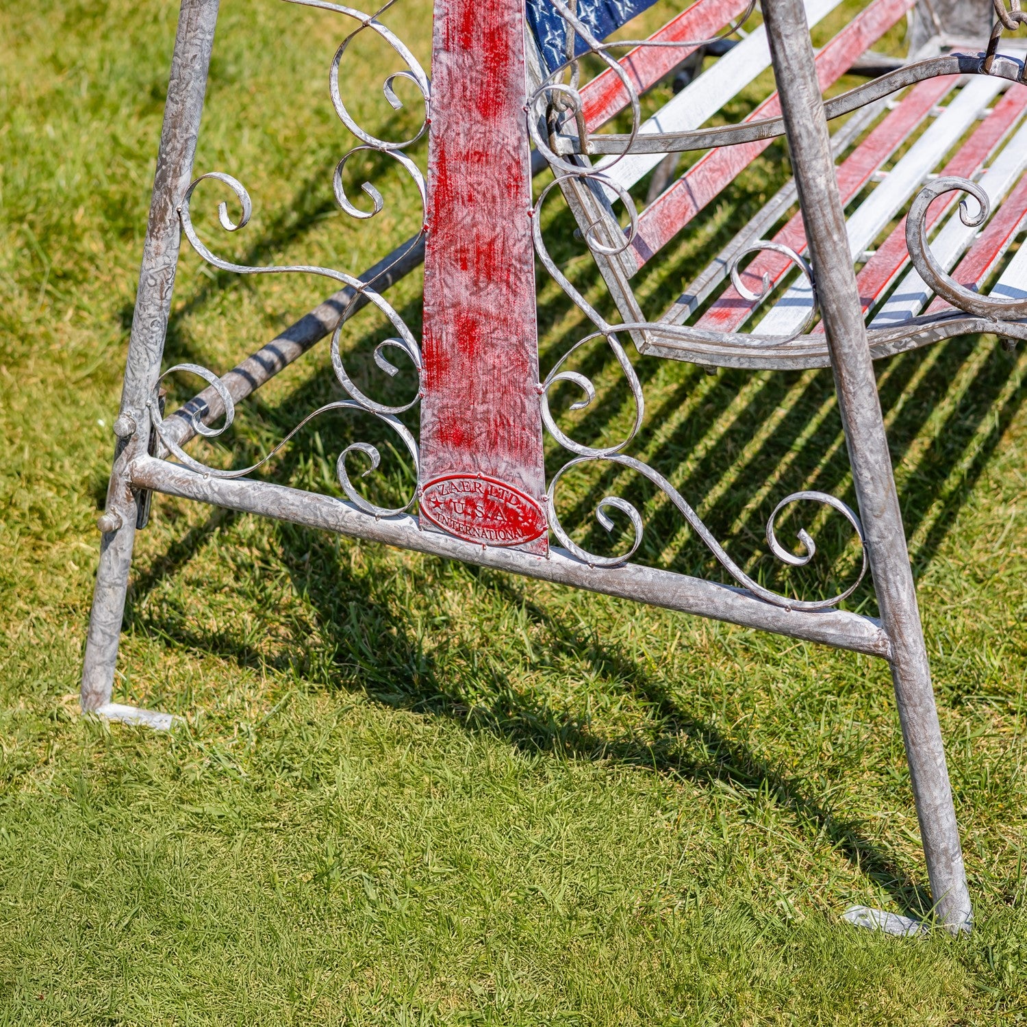 A close-up outdoor shot of a weathered metal garden bench on green grass. The bench has ornate scrollwork on the frame, a distressed red vertical support, and a red, white, and blue striped slatted seat and backrest.