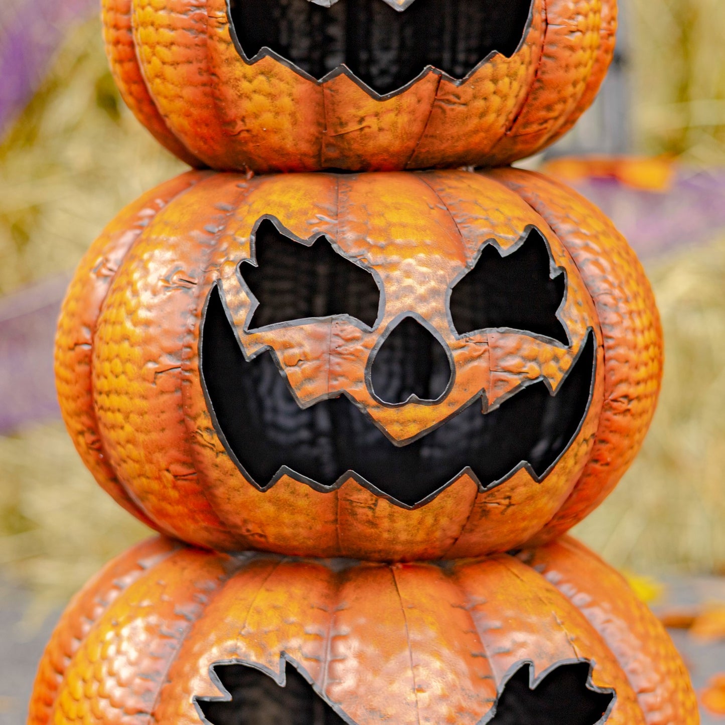 Three stacked orange jack-o'-lantern pumpkins with carved black faces, photographed close up against a softly blurred autumnal background.