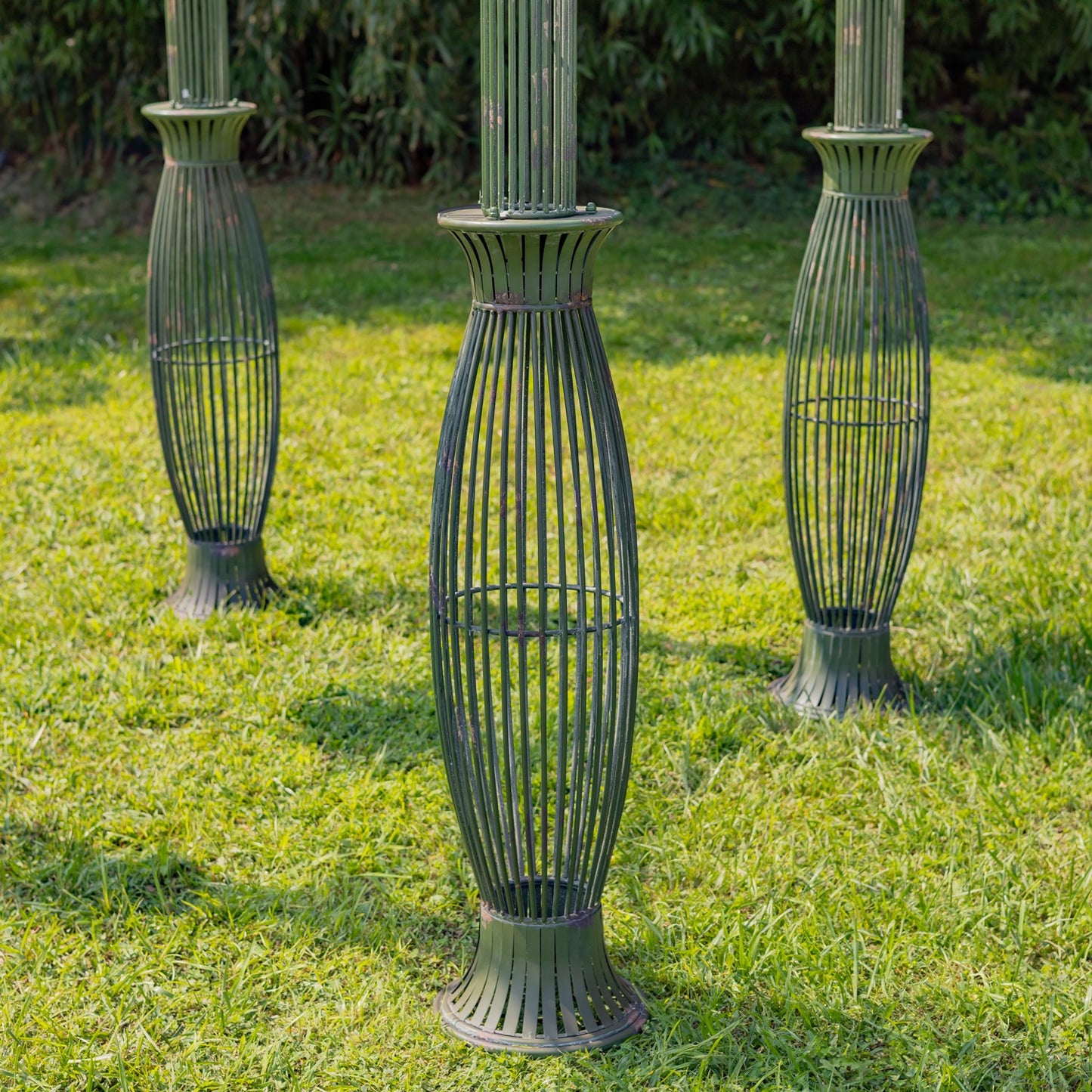 Three dark metal garden column guards with a ribbed, vase-like design standing on a green grass lawn.