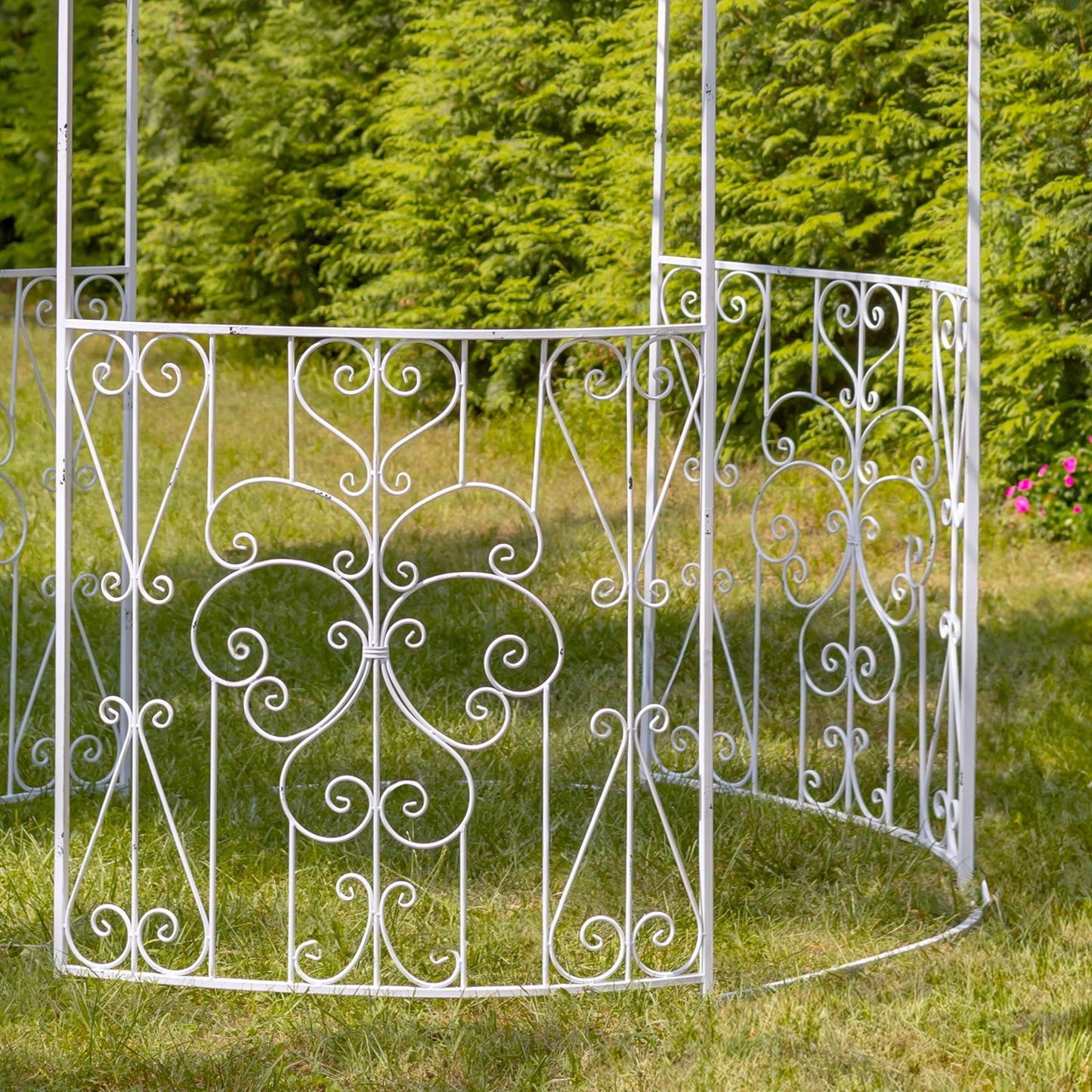 White wrought iron garden gazebo with ornate scrollwork, set on a grassy lawn with a green hedge background.