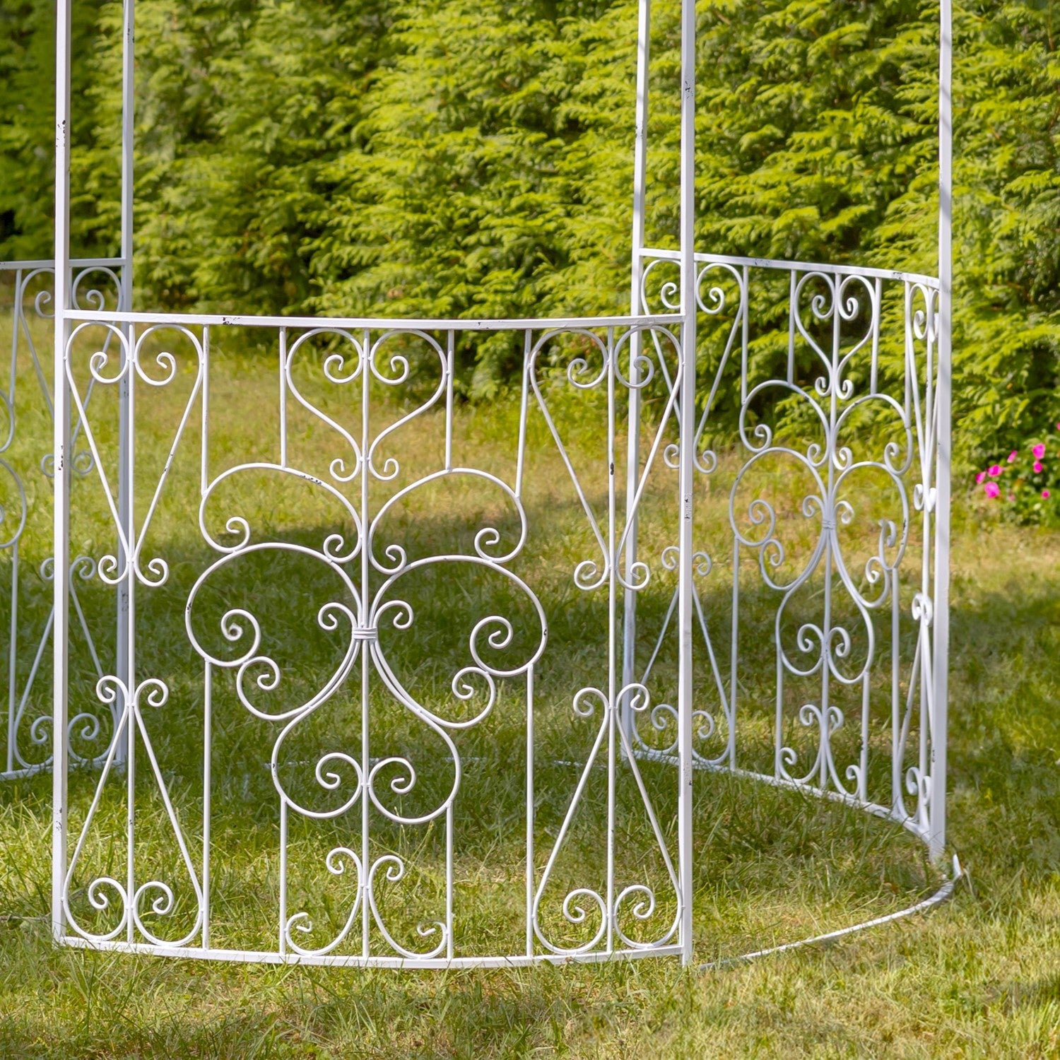 White wrought iron garden gazebo with ornate scrollwork, set on a grassy lawn with a green hedge background.