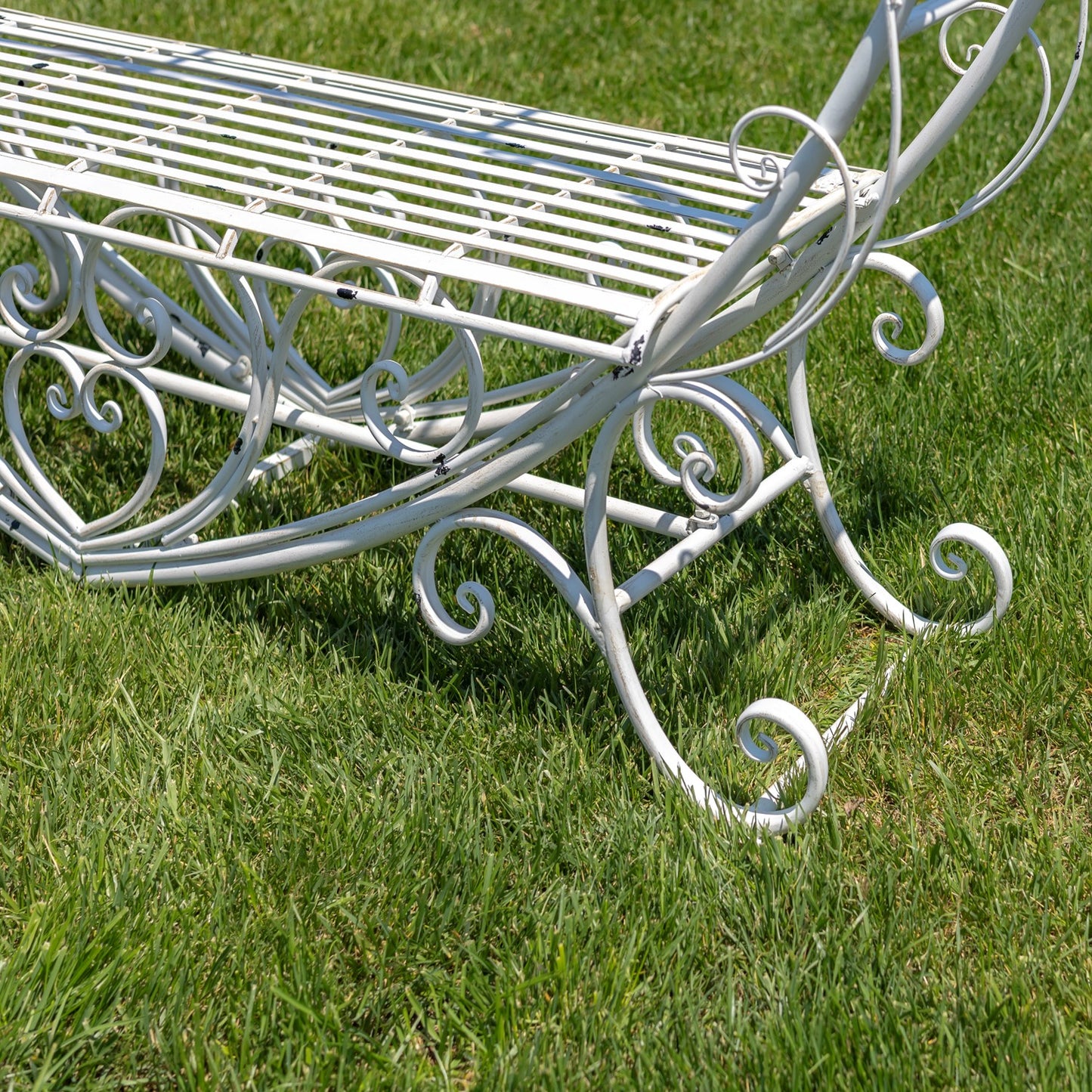 A white wrought iron garden bench with ornate scrollwork details, shown on a green grass lawn.