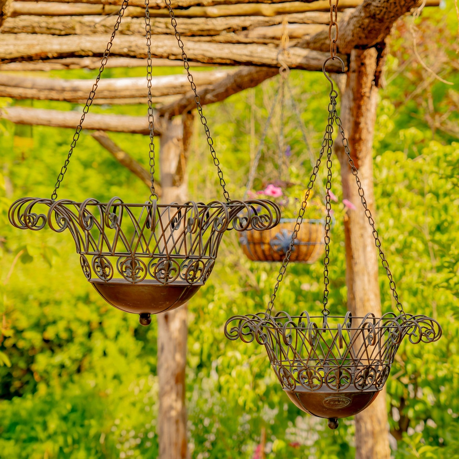 Two ornate metal hanging planters with scrollwork tops and bowl-shaped bases, suspended by chains from a rustic wooden pergola in a garden setting.