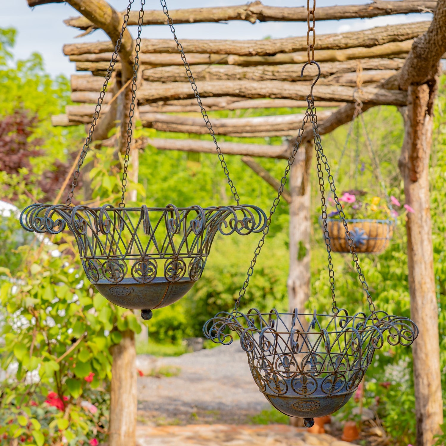 Two ornate hanging metal planters with scrollwork details and chain suspensions, displayed outdoors under a rustic wooden trellis with green garden foliage in the background.