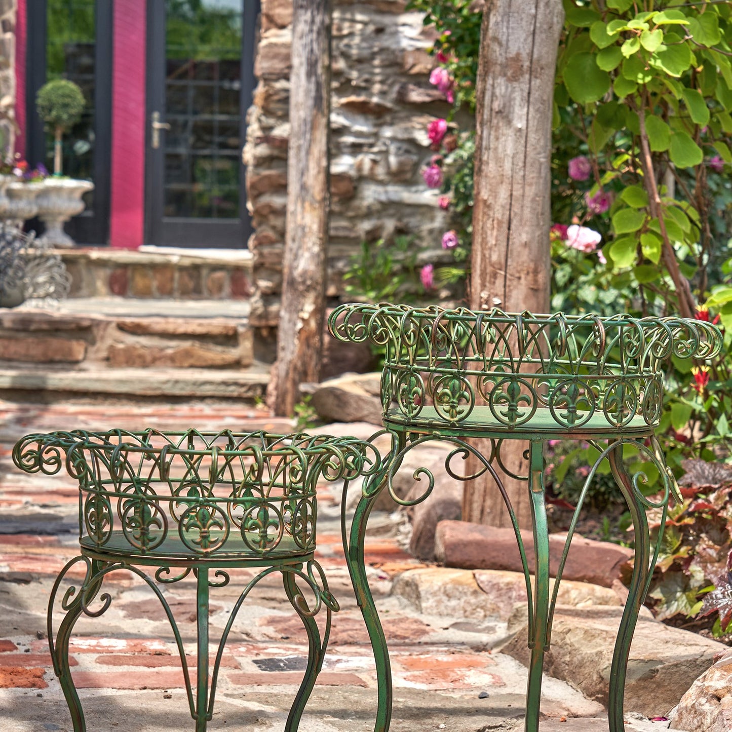 Two ornate green metal garden planters or small tables with decorative scrollwork, placed outdoors on a stone patio in front of a garden doorway.