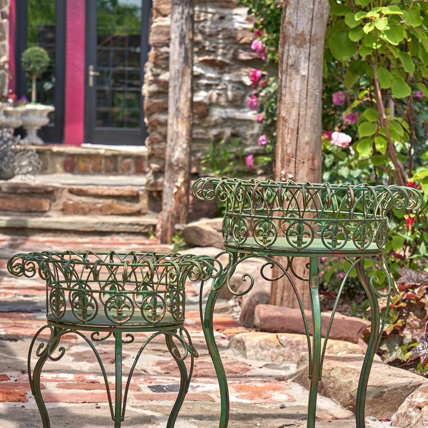 Two ornate green metal garden planters or small tables with decorative scrollwork, placed outdoors on a stone patio in front of a garden doorway.