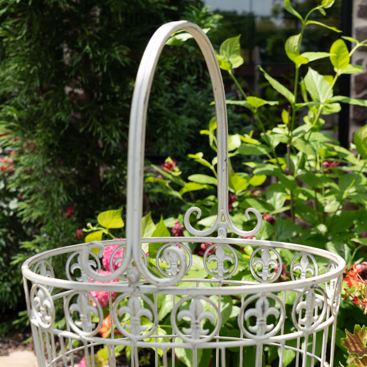 A white ornate metal garden basket with a tall arched handle, set against a lush green garden background with foliage and small pink flowers.