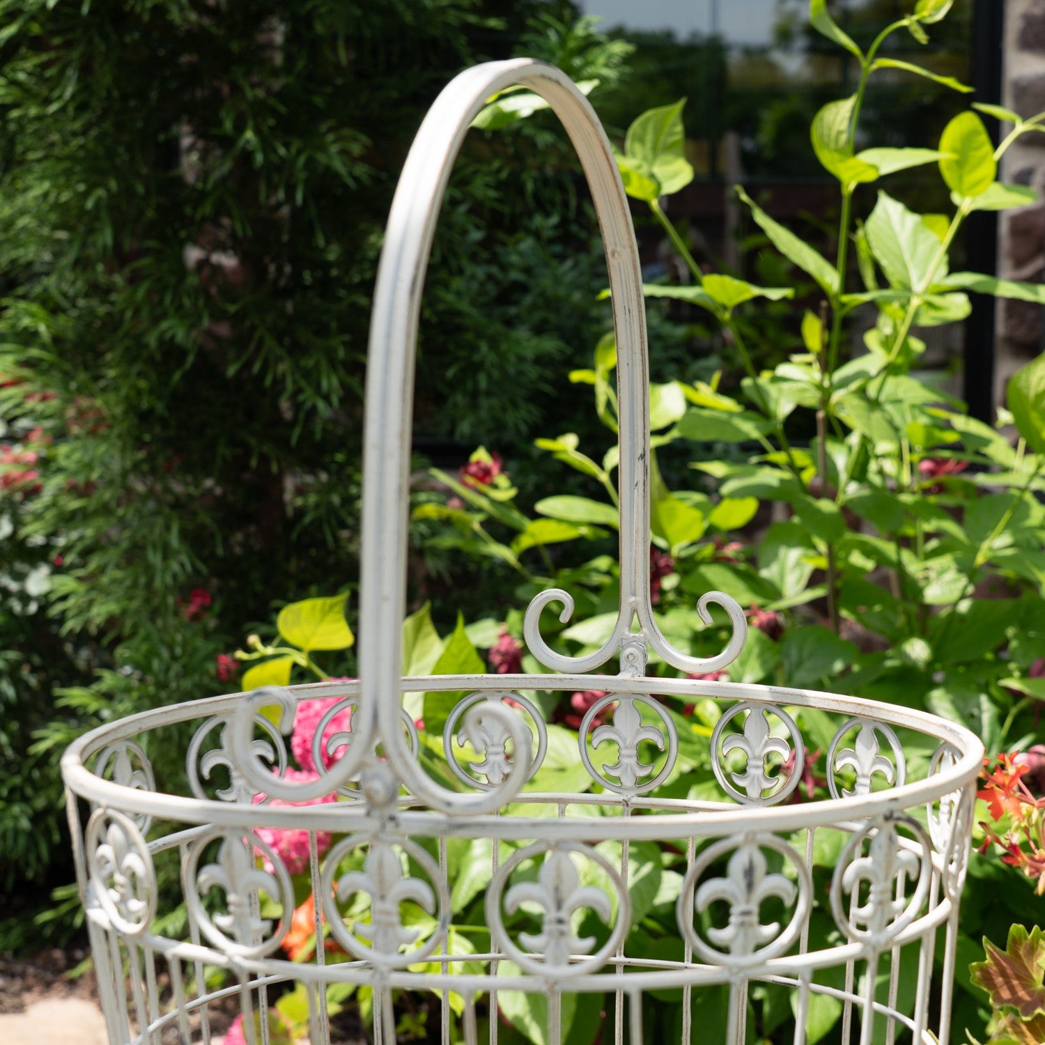 A white ornate metal garden basket with a tall arched handle, set against a lush green garden background with foliage and small pink flowers.