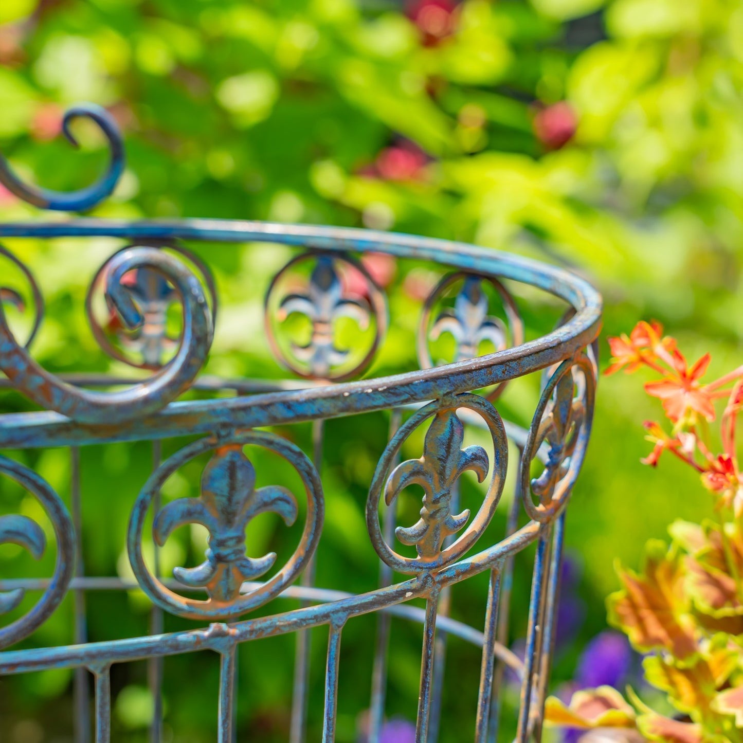 A close-up of a decorative metal garden stake or planter frame with a weathered blue and rusted bronze finish, featuring circular scrollwork and fleur-de-lis motifs, set against a blurred green garden background with flowers.