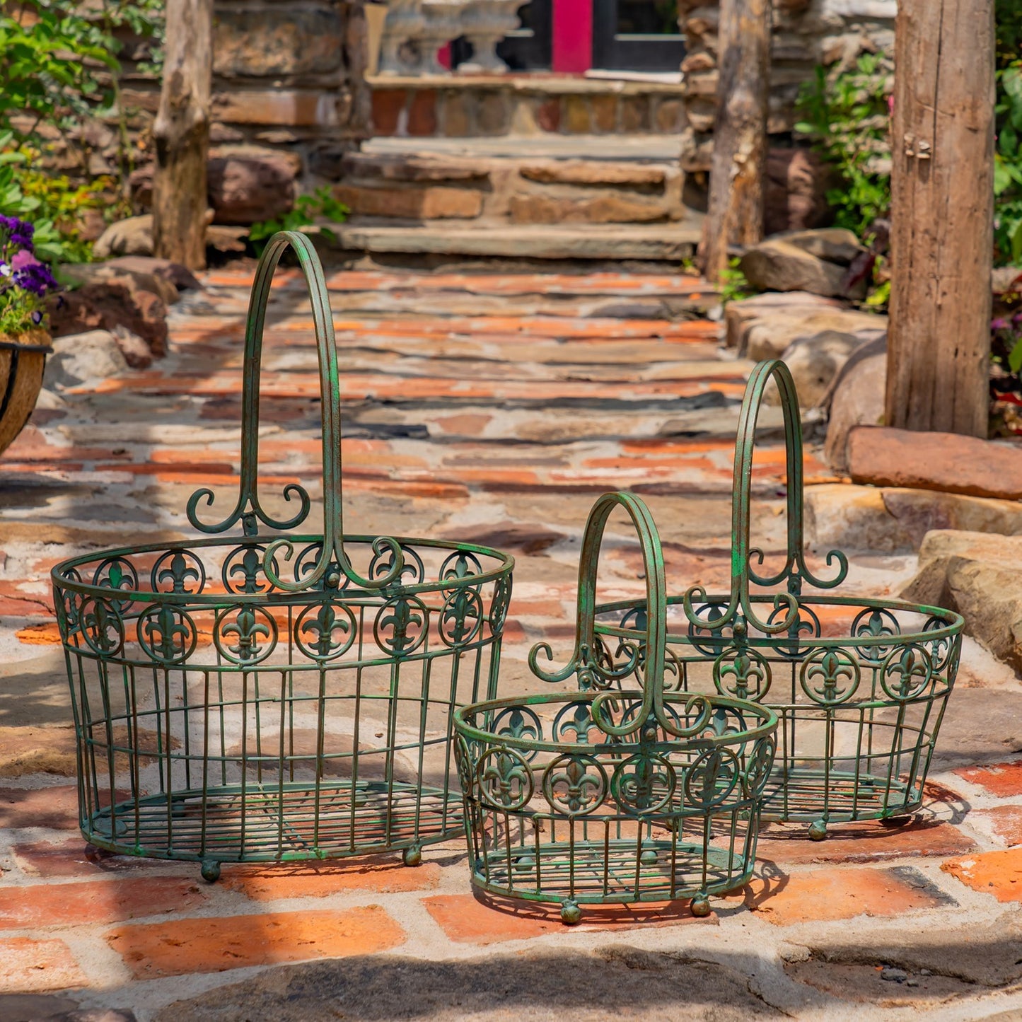 Three green metal wire baskets with decorative scrollwork and arched handles, arranged on a stone patio in an outdoor garden setting.