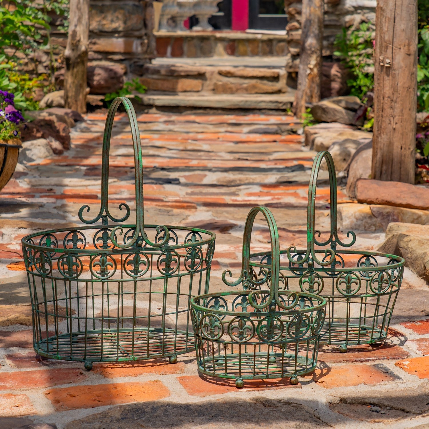 Three green metal wire baskets with decorative scrollwork and arched handles, arranged on a stone patio in an outdoor garden setting.