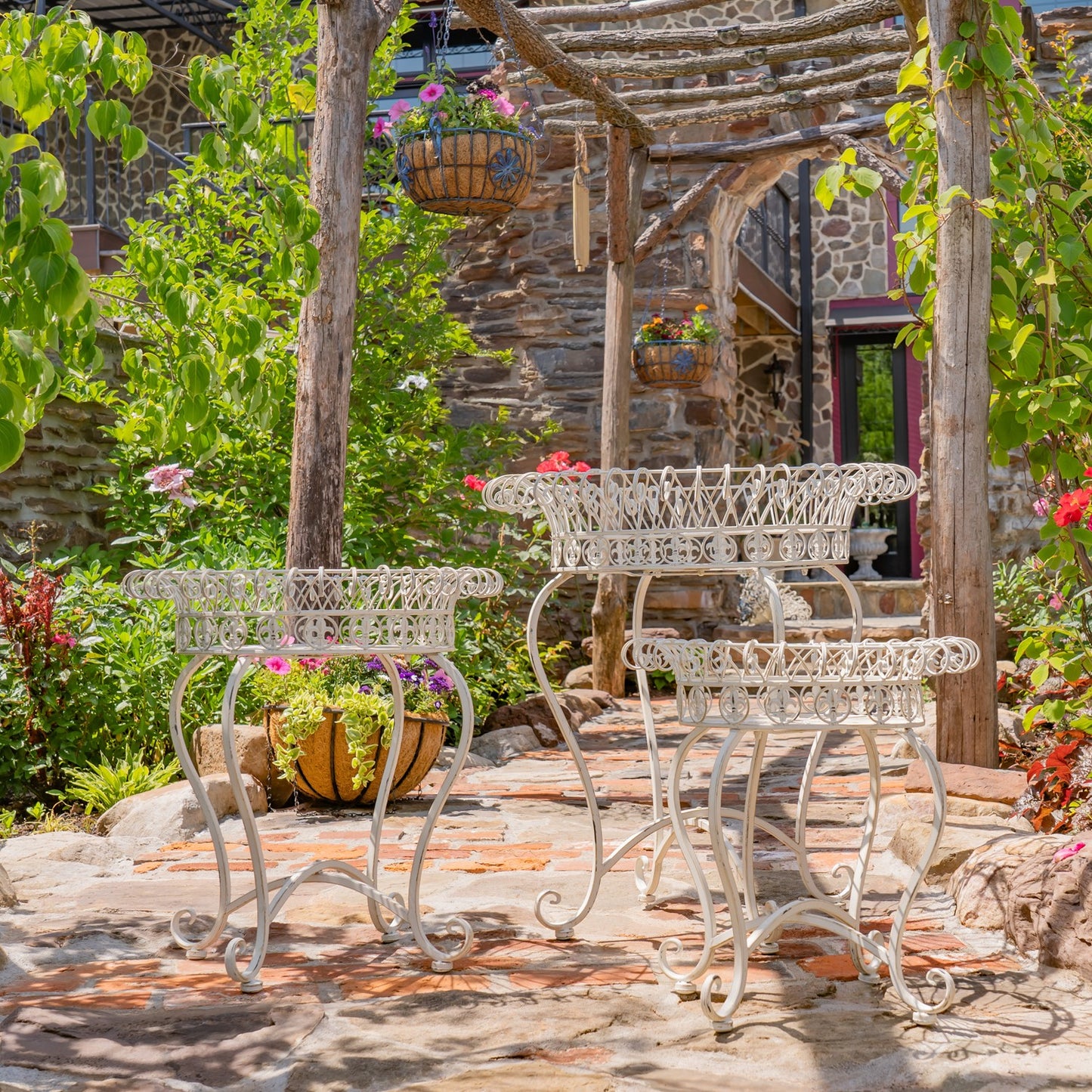 Three white wrought-iron garden tables with matching planters arranged on a stone patio under a rustic wooden pergola with climbing vines and potted flowers.