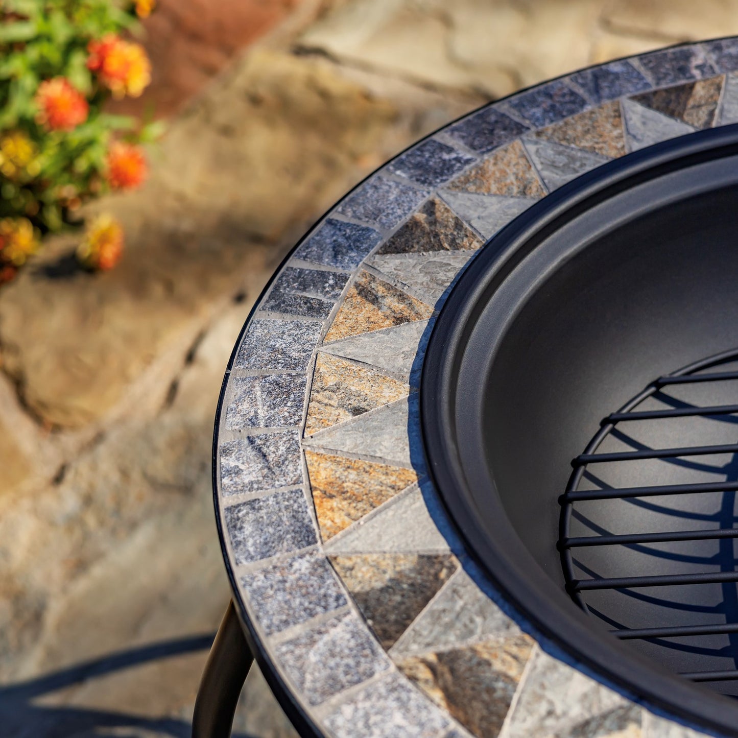 Close-up of a round outdoor fire pit table with a mosaic stone tile rim and a black metal bowl interior, photographed outdoors on a stone surface with flowers blurred in the background.