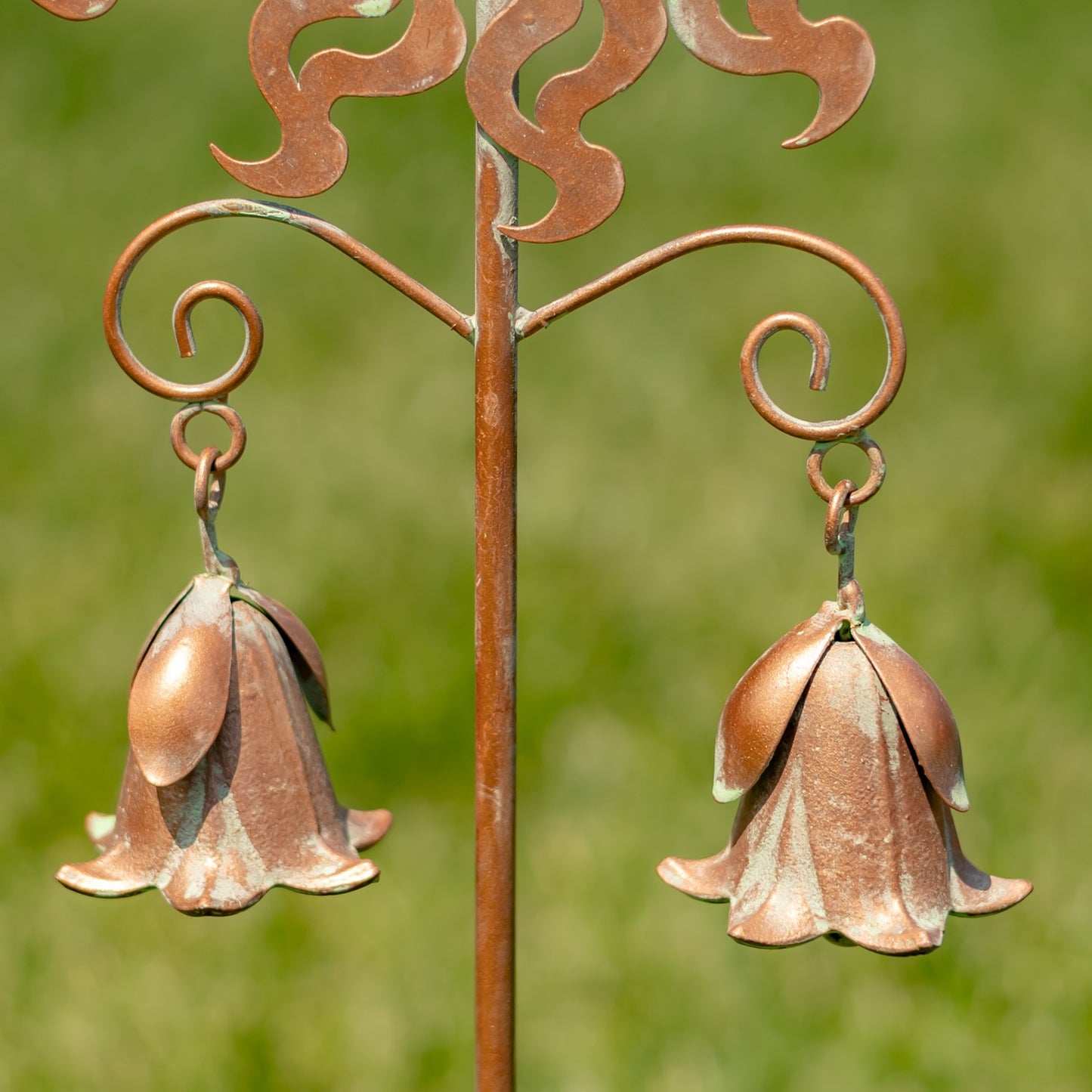 A copper-toned metal garden stake with two hanging bell-shaped flower ornaments, photographed outdoors against a blurred green grass background.