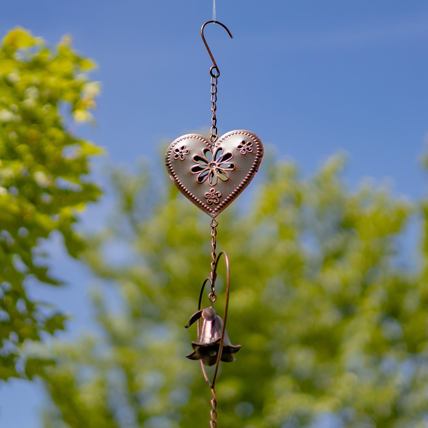 A copper-toned metal garden wind spinner featuring a bell-shaped flower suspended inside a heart-shaped wire frame, set against a blurred green grass background.