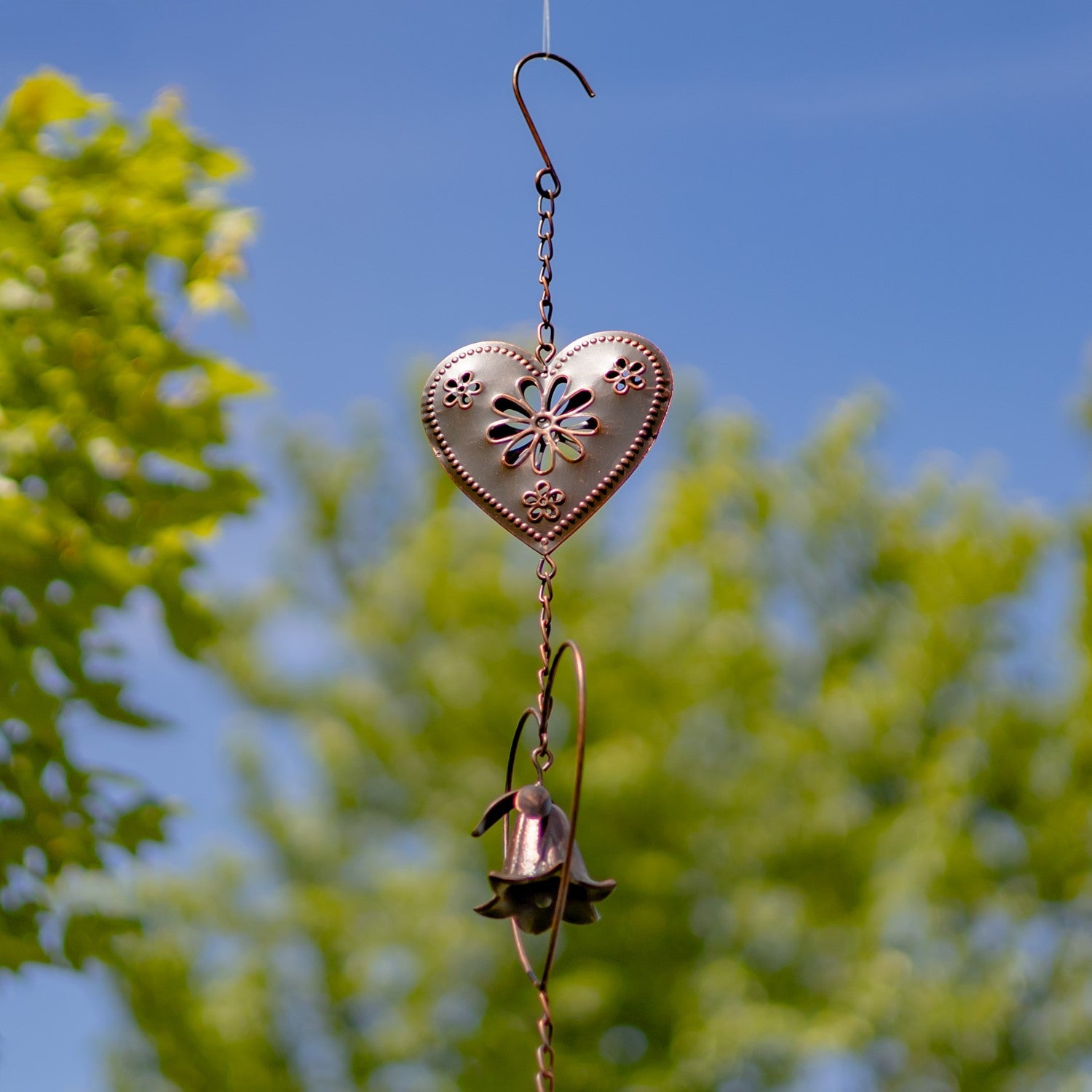A copper-toned metal garden wind spinner featuring a bell-shaped flower suspended inside a heart-shaped wire frame, set against a blurred green grass background.