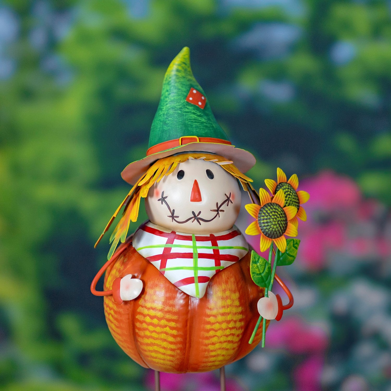 A decorative autumn scarecrow figure made from a pumpkin body, green hat, straw hair, stitched face, and sunflower accessory, photographed against a blurred green garden background.